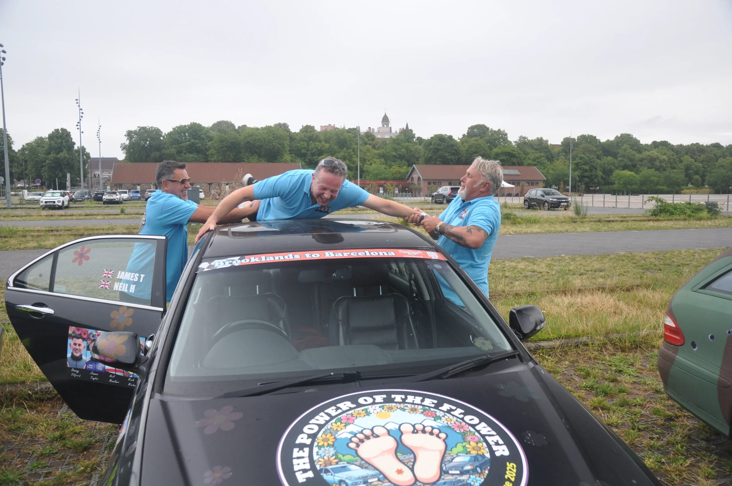 Three men in blue shirts celebrating around a decorated black car, one man lying on the car roof, two smiling men holding his arms, in an outdoor parking lot with trees and buildings in the background.