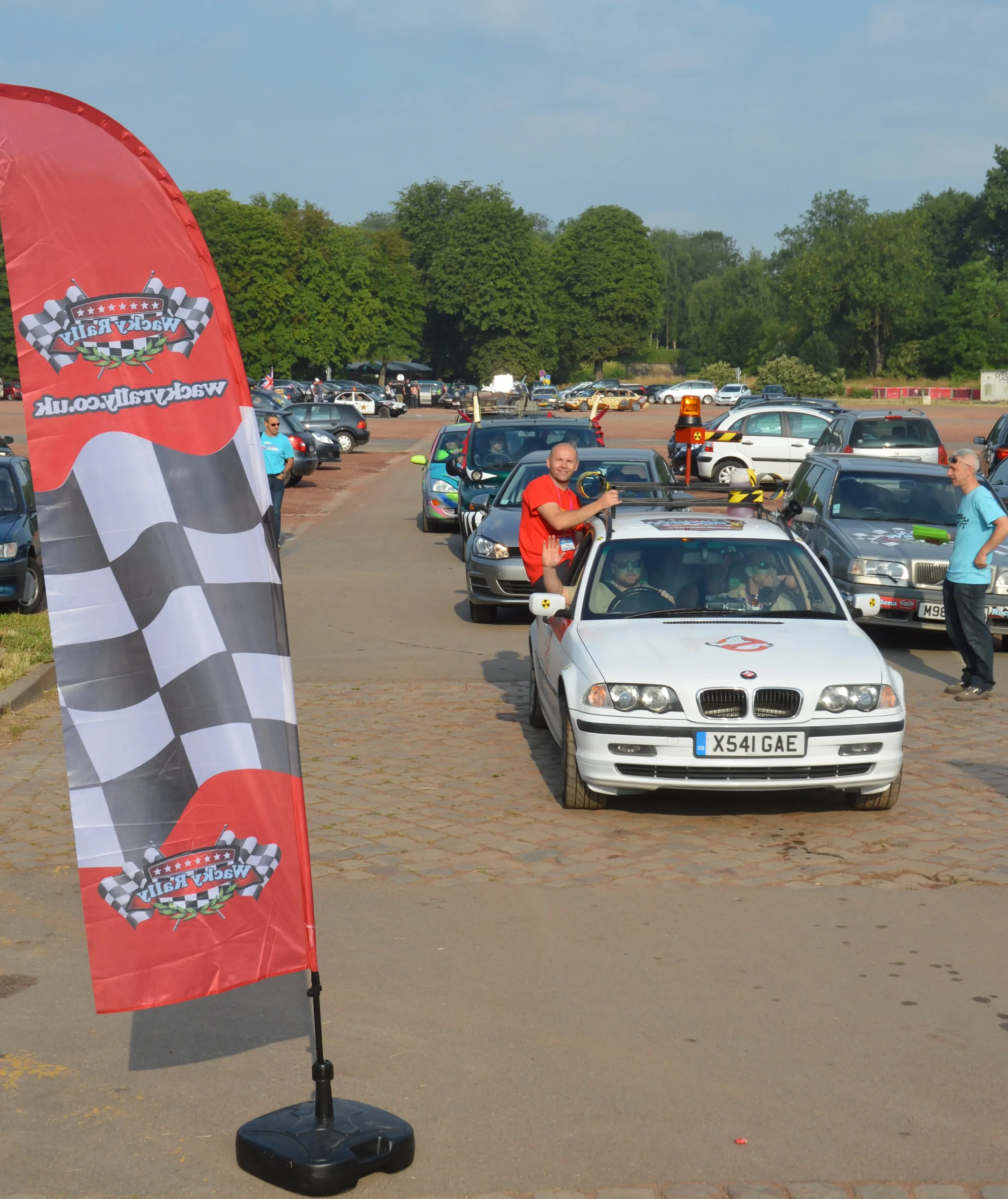 Cars lined up at a motorsport event with a racing flag banner on the left and people standing around, some in racing gear.