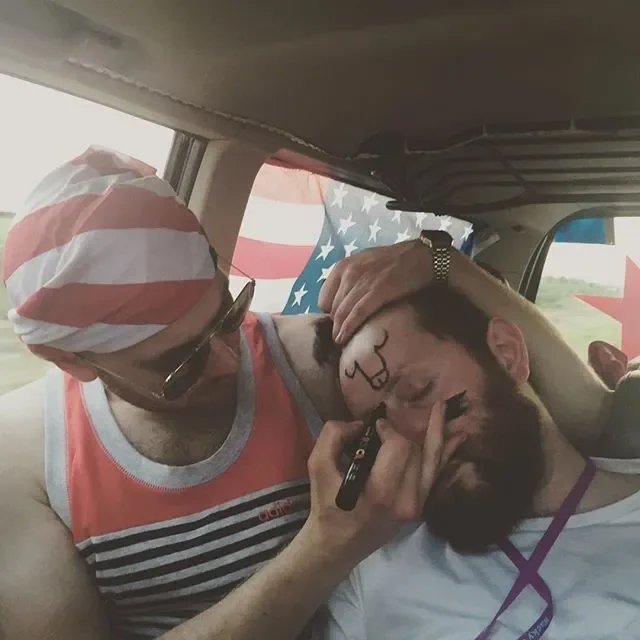 A woman with a bandana and sunglasses is painting the face of a man with a beard, who appears to be lying down in a vehicle. The woman is wearing a sleeveless striped tank top and is holding makeup brushes. The man is dressed in a white shirt and has