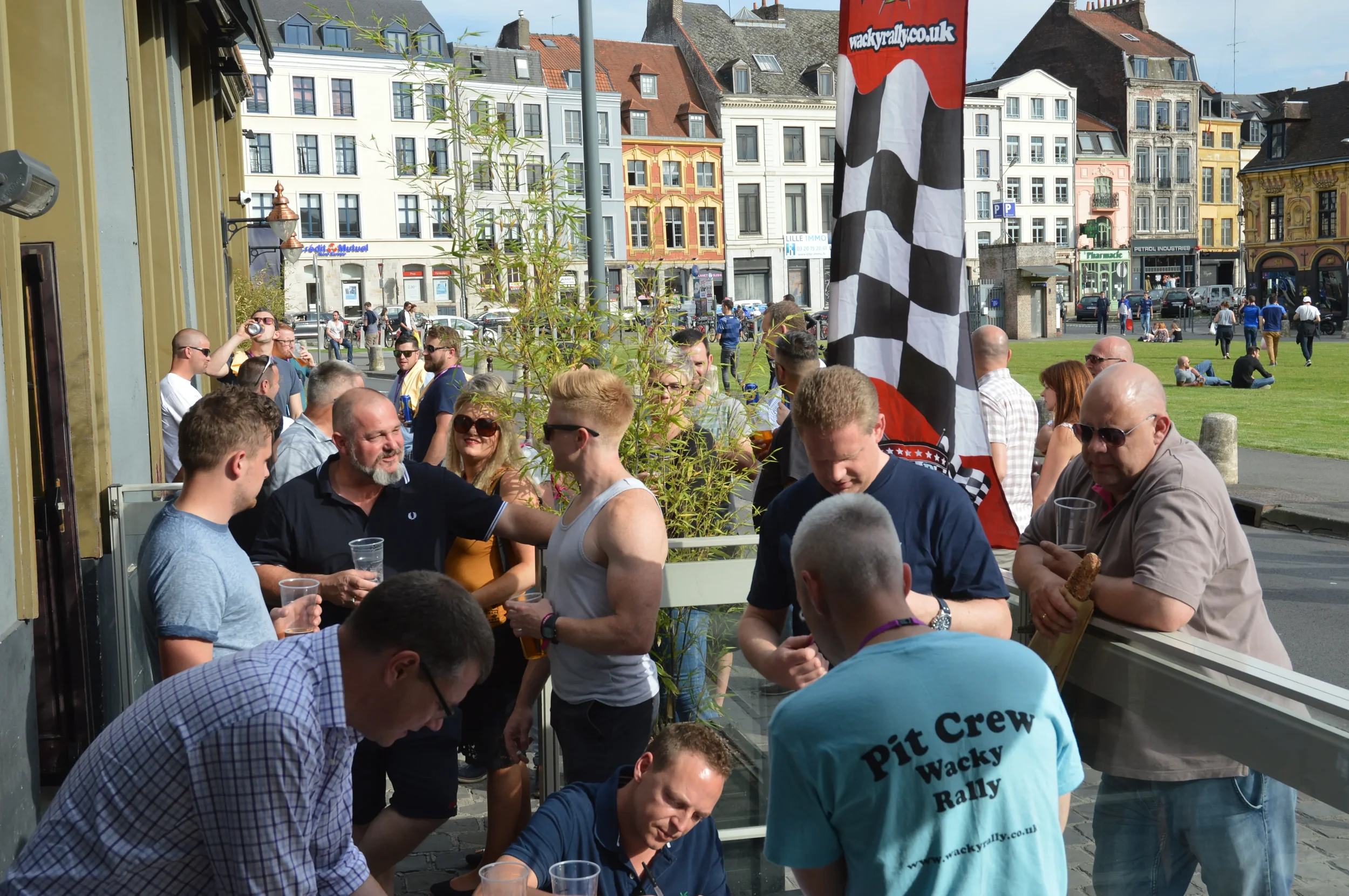 A group of people gathered outside a building, some holding drinks, with a large checkered flag and colorful buildings in the background, indicating a casual social event or celebration.