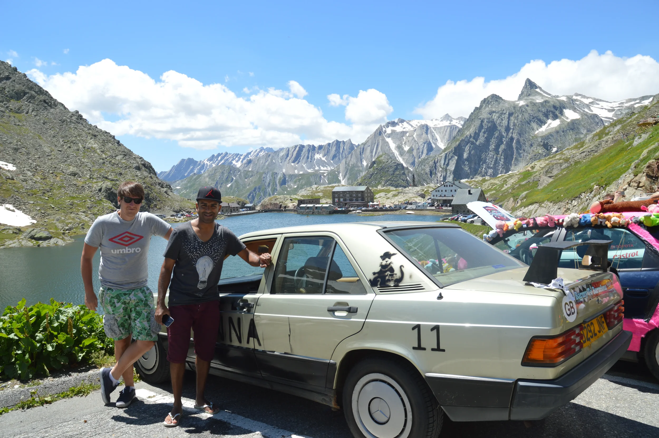 Two men standing next to a vintage car with mountainous scenery and a lake in the background.