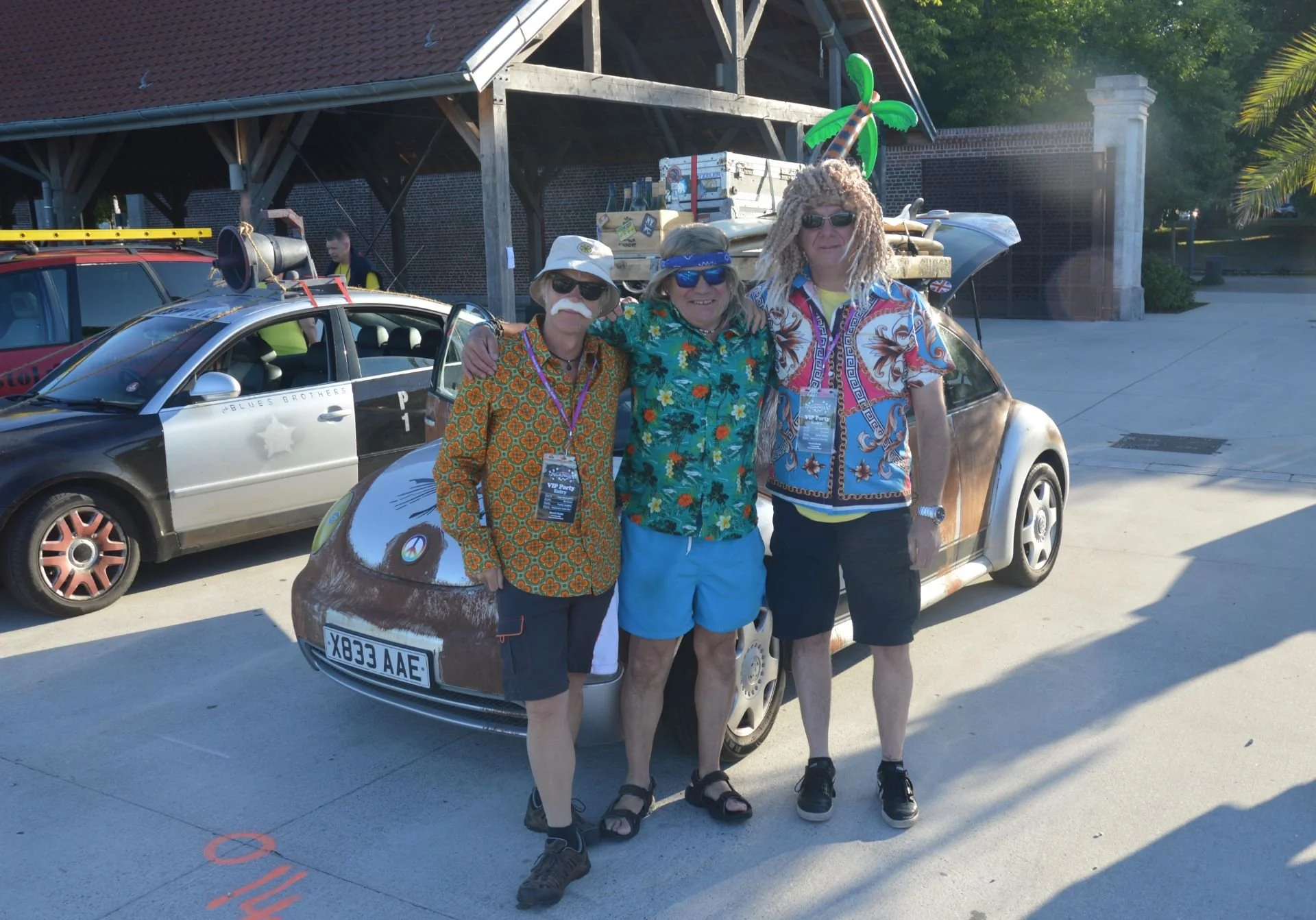 Three men dressed in colorful, retro outfits standing together outdoors, with a themed car decorated with tropical and music-related items in the background.