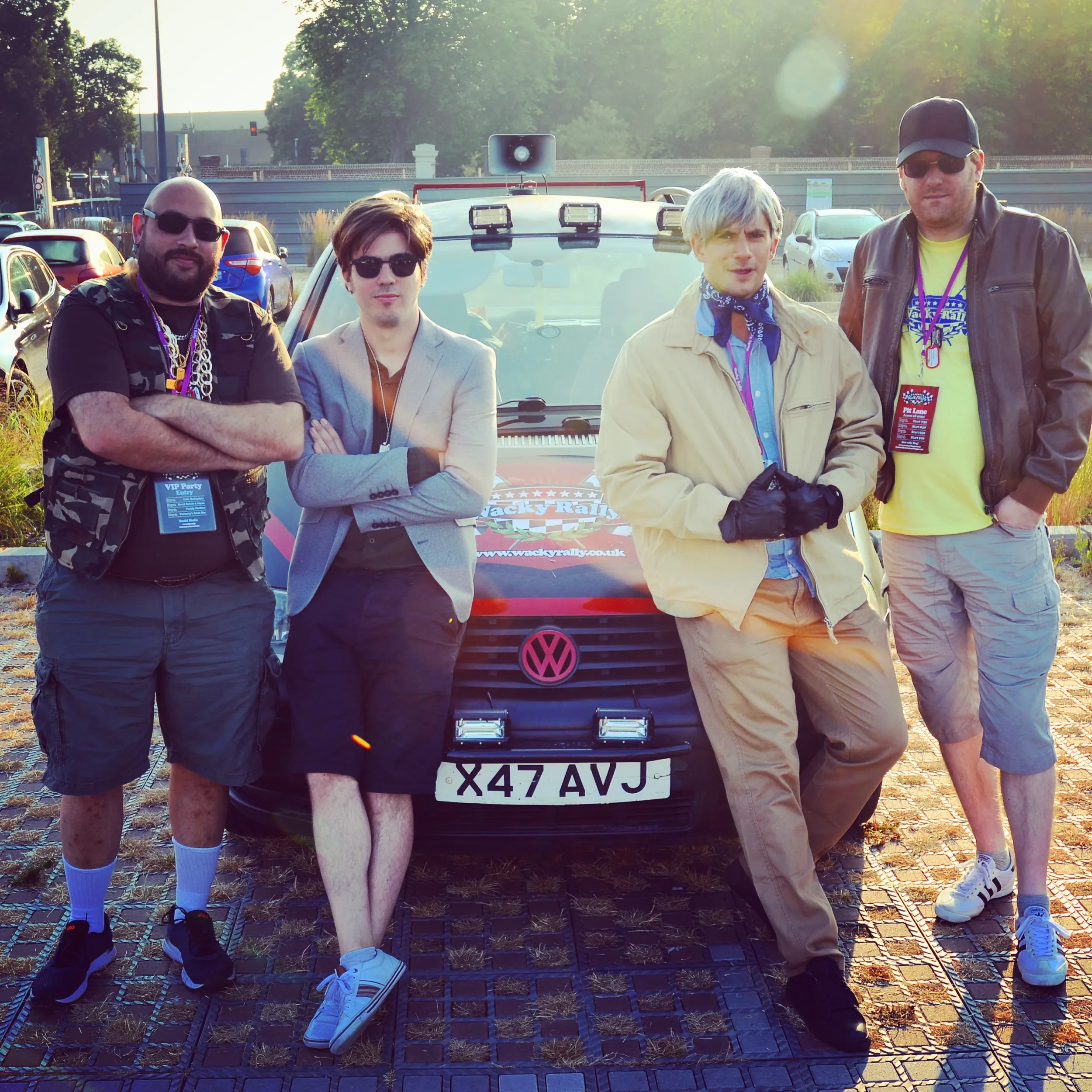Four men standing in front of a black Volkswagen car with a license plate X47 AVJ, outdoors on a paved area during daytime, with trees and parked cars in the background. The men are posing for the photo, some with crossed arms, dressed casually, and 