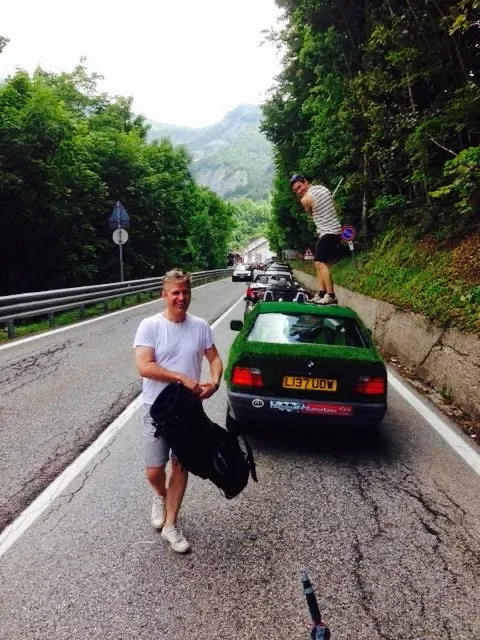 Two people with a small green car on a mountain road; one sitting on the roadside, the other walking toward the camera, holding a jacket.