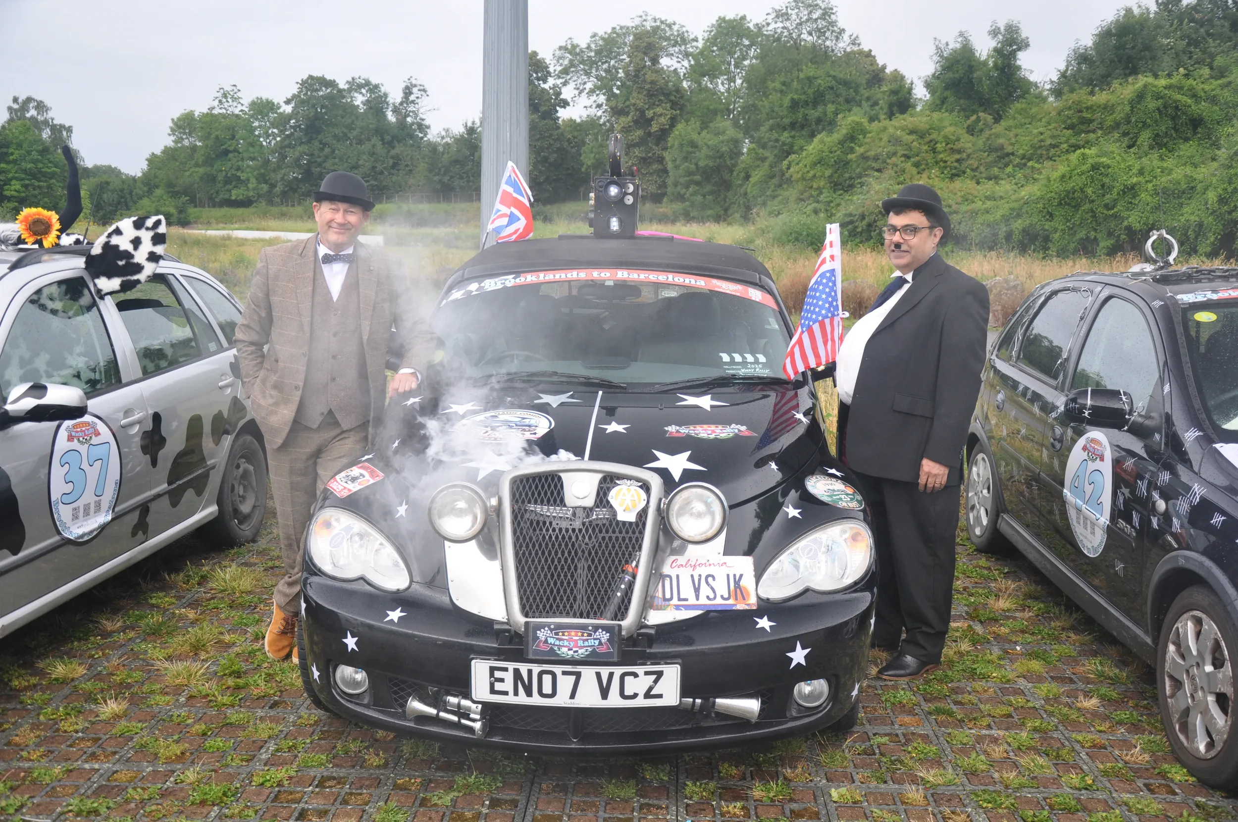 Two men dressed in vintage-style suits and hats standing next to a creatively decorated black car with American flags, stars, and racing numbers, during an outdoor event.