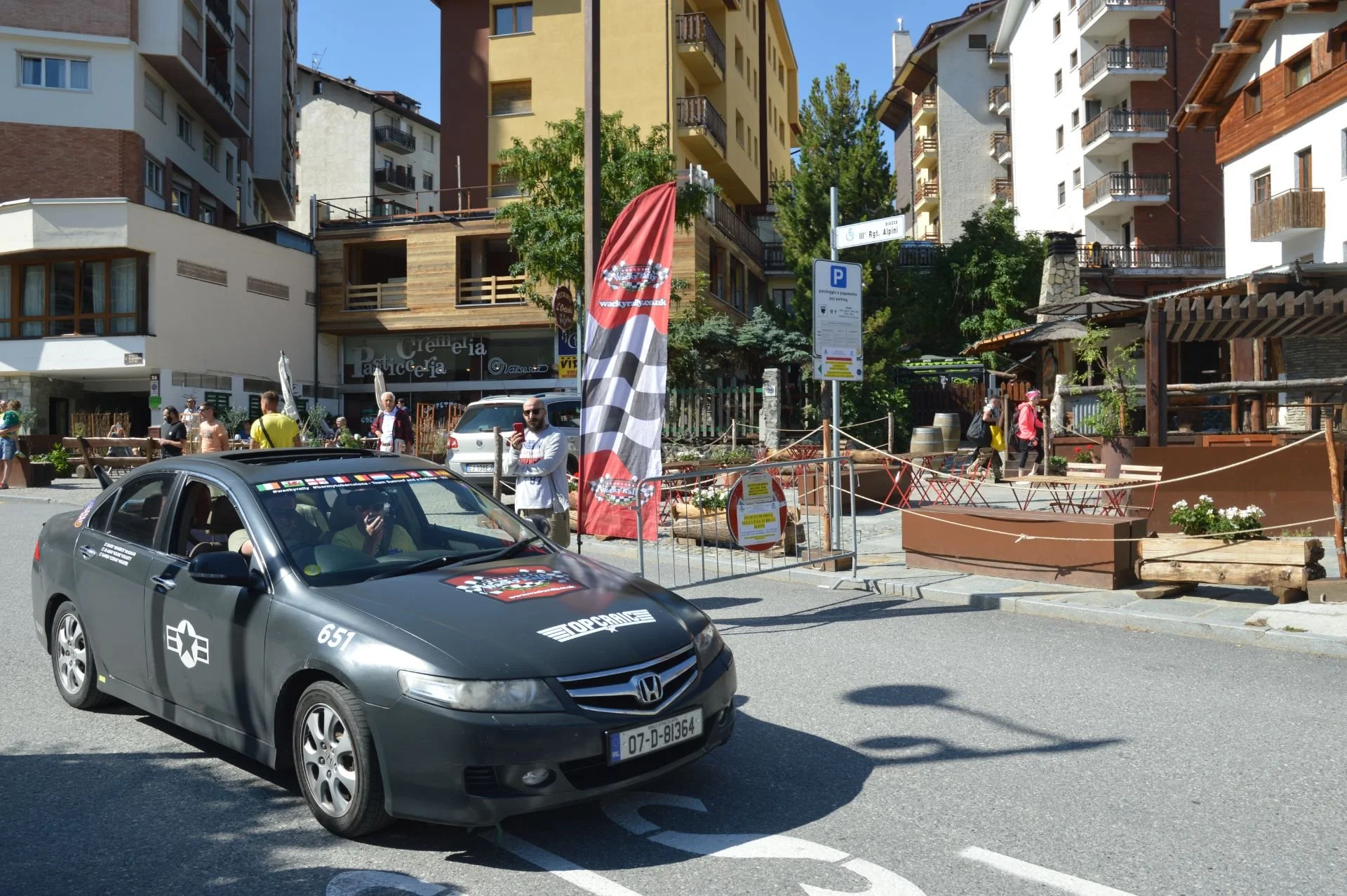 A black Honda police car with racing decals parked on a city street, with people walking and standing nearby, colorful buildings in the background, and a red flag with checkered patterns.