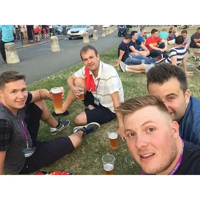 Group of young men sitting on the grass with drinks, smiling for a selfie during an outdoor event, with more people sitting on the grass in the background.