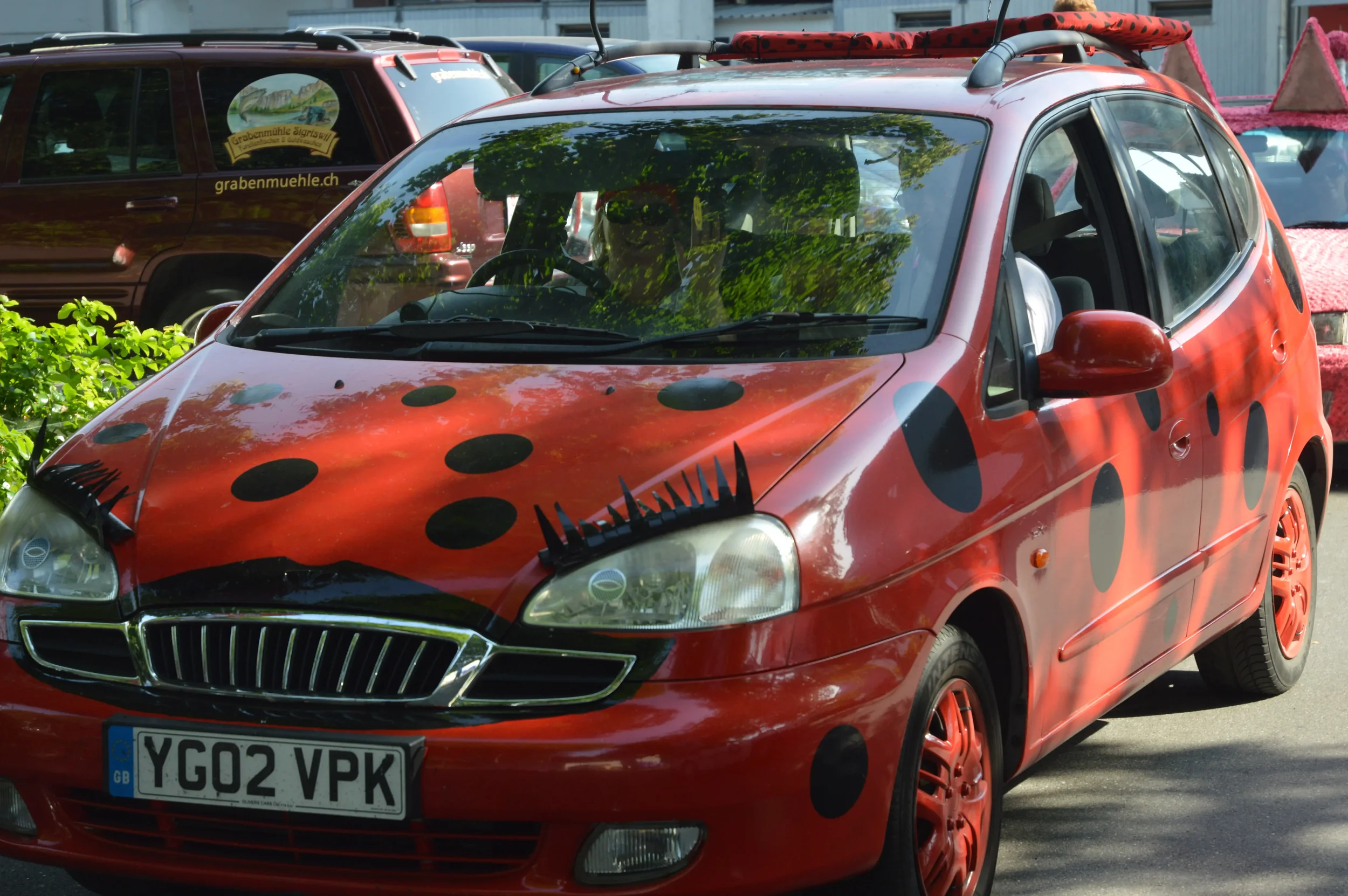 A red car decorated with black polka dots resembling a ladybug, with a surfboard on top and eyelashes on the headlights.