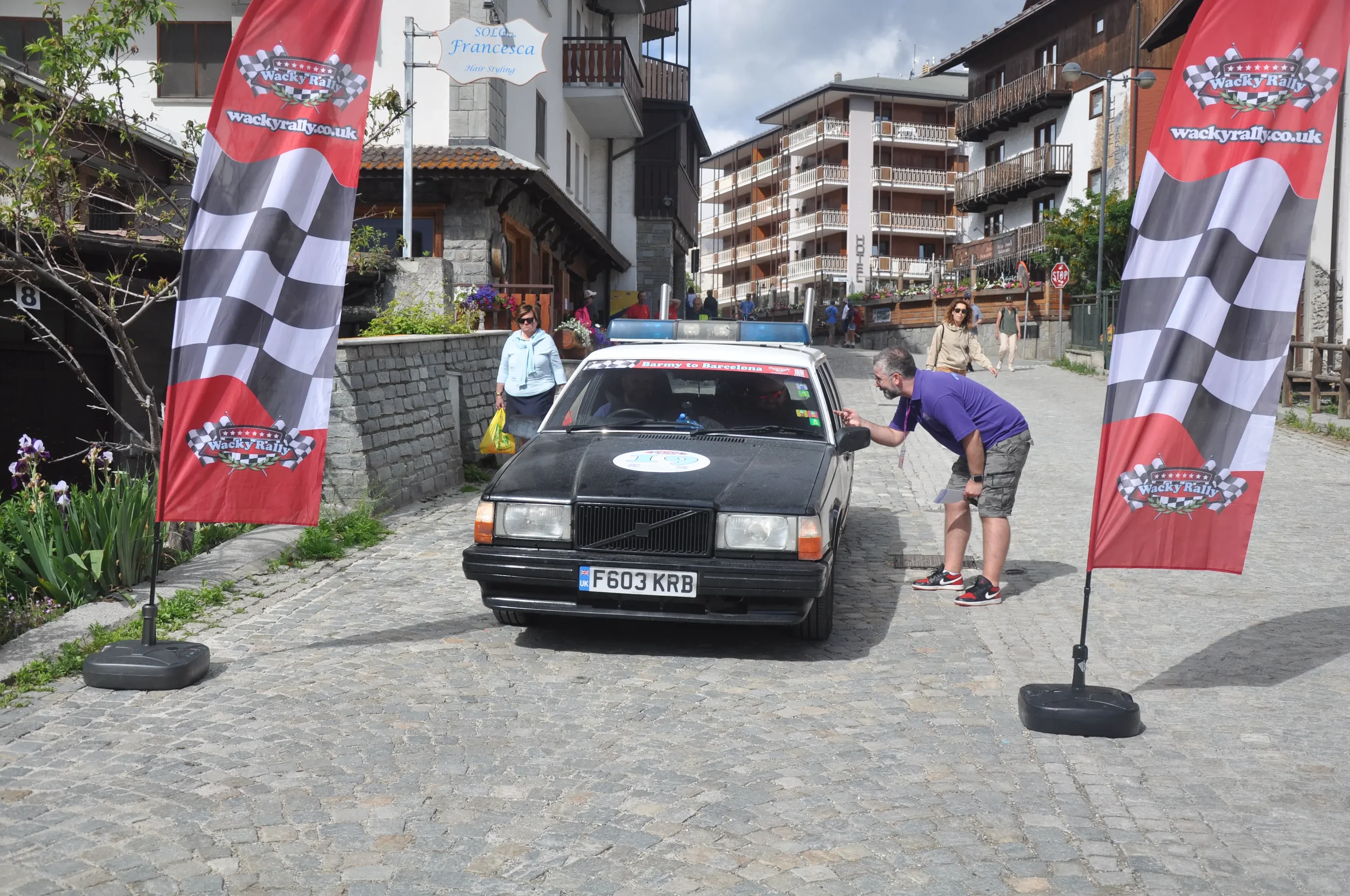 A man is leaning into a black vintage Volvo car at a rally event, with flags displaying the Wacky Rally logo on either side.