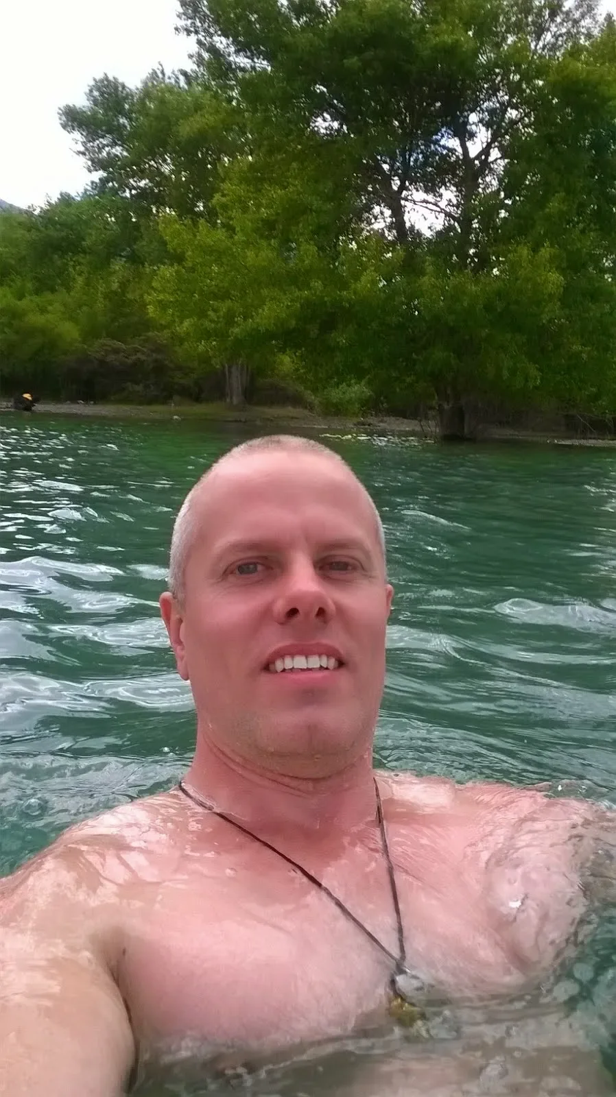 A shirtless man with a necklace taking a selfie in a lake with green water, surrounded by trees and greenery.