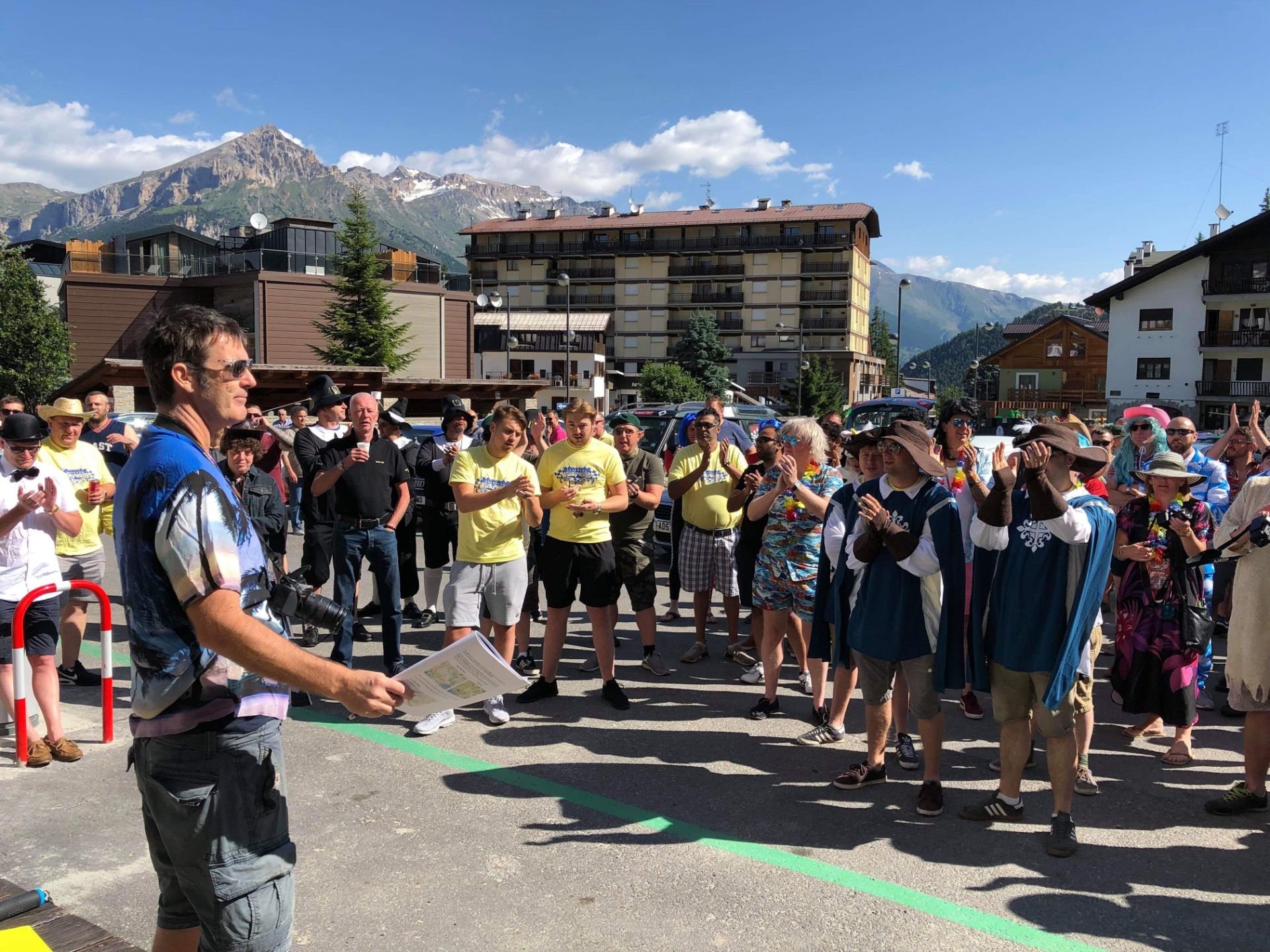 A group of people gathered outdoors in a city square, listening to a man with sunglasses holding a paper, with mountains and buildings in the background on a sunny day.