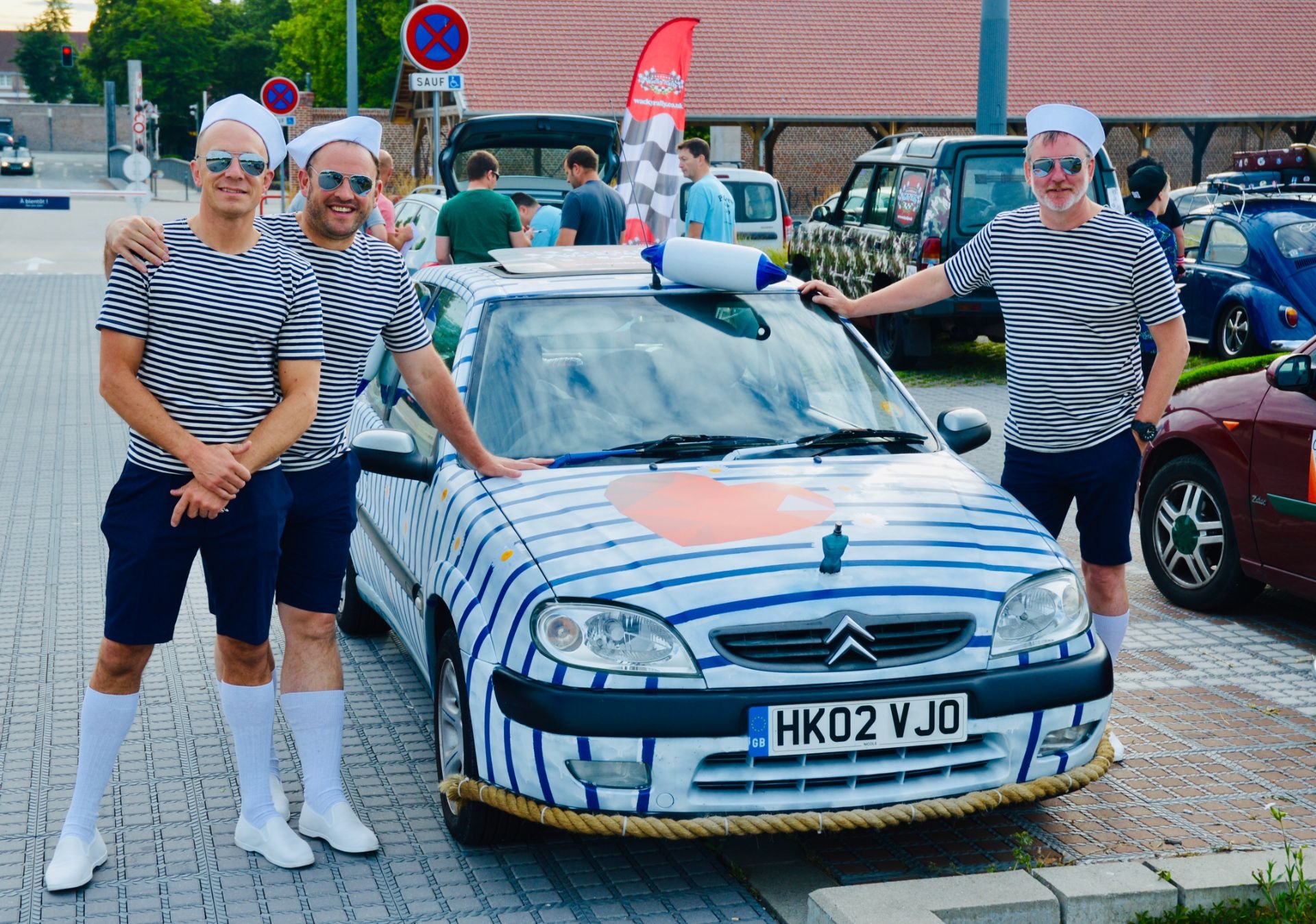 Three men dressed in matching striped shirts, shorts, white socks, and sailor hats standing next to a small decorated car with a large salt shaker on top. The car is adorned with blue and white stripes and a rope around the front bumper. They are out