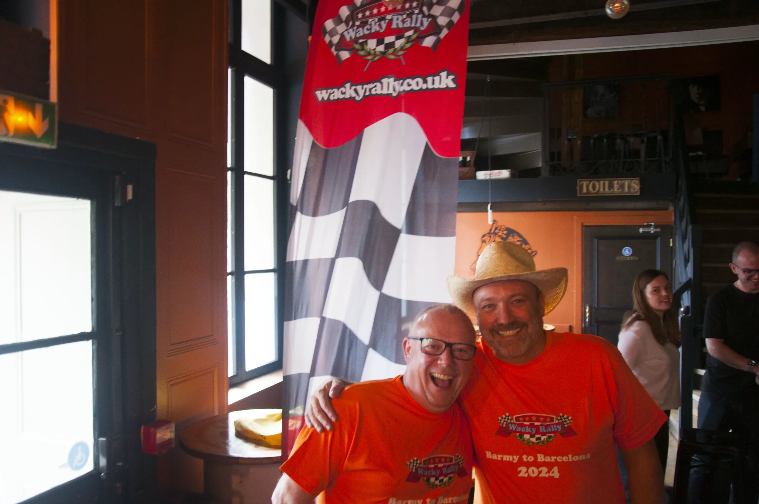 Two men wearing orange shirts and a straw hat smiling and posing together inside a room with a checkered flag banner behind them.