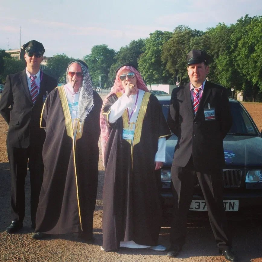 Four individuals in formal attire, two in academic robes and others in suits, standing outdoors near a black car with trees in the background.