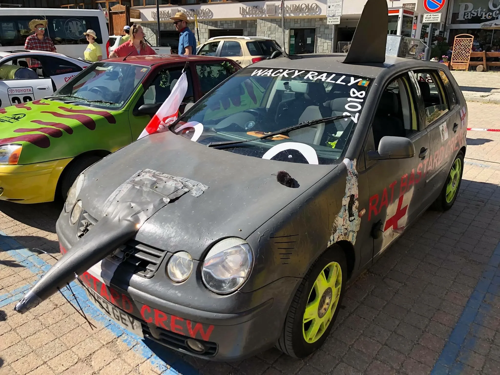A car decorated for a rally race with various stickers and painted details, resembling a makeshift race vehicle, parked among other cars and people in a sunny outdoor area.