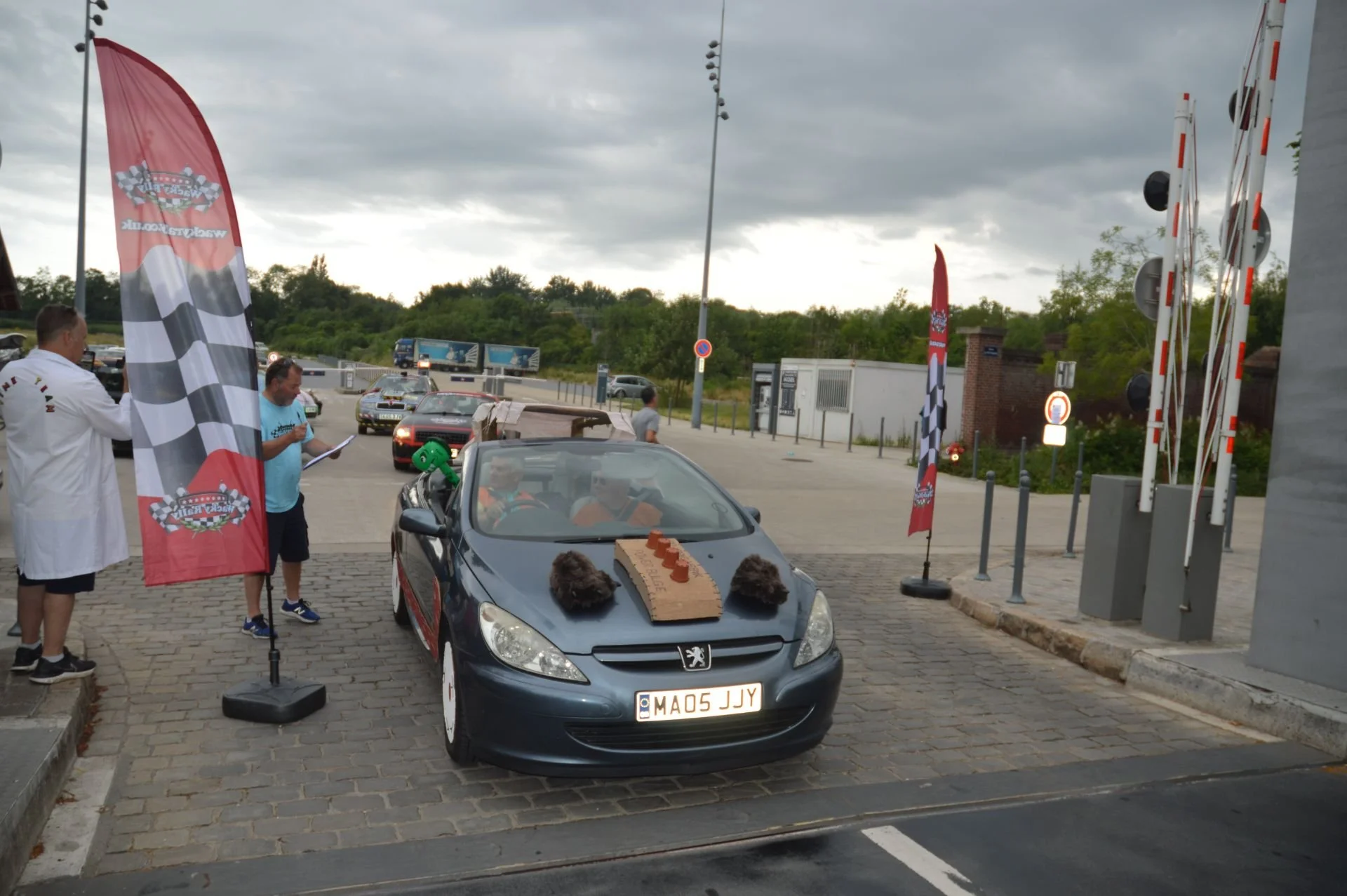 Car decorated with racing-themed items, including foam rocks and a brake pad, parked near flags and barriers at a racing event, with people gathered around.