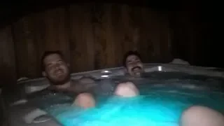 Two men relaxing in a hot tub or jacuzzi, partially submerged in water, with wooden paneling in the background.