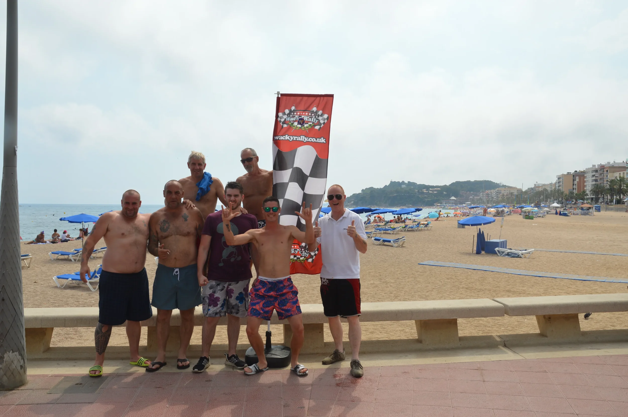 Group of eight men on a beach promenade in swimwear, posing in front of a Wacky Rally flag, with lounge chairs and umbrellas on the sandy beach and buildings in the background.