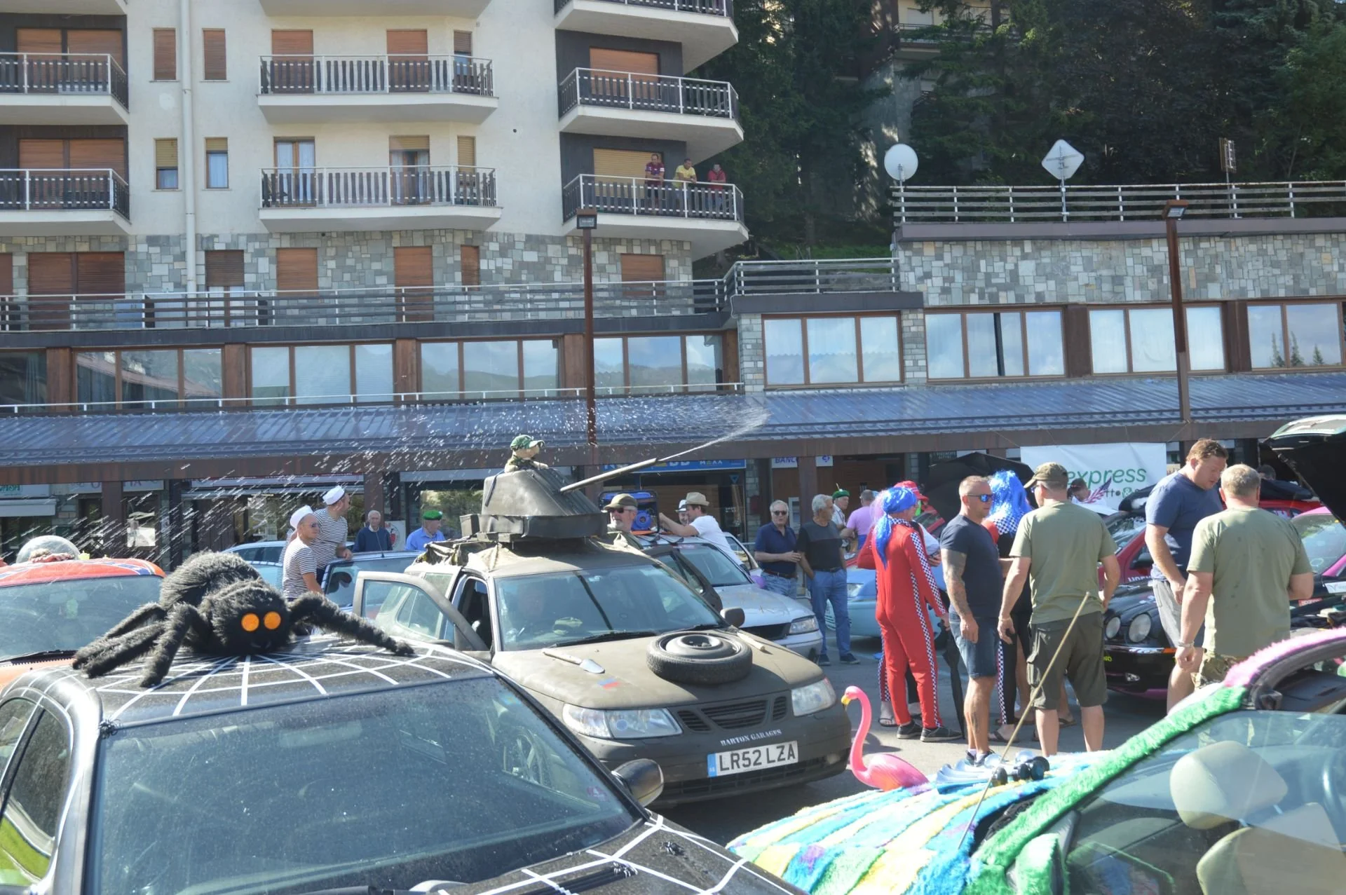 People gathered in a parking lot decorated for a celebration, with some dressed in costumes. Several cars are decorated with various props, and a tank is present with water being sprayed. People are standing and socializing, with a multi-story buildi