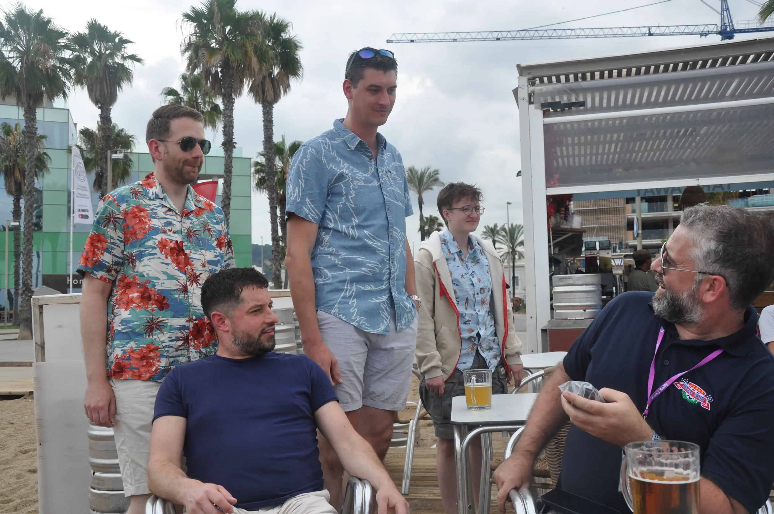 Group of five people socializing outdoors near a beach with palm trees, drinks on tables, and a weathered building in the background, some wearing tropical shirts and sunglasses.