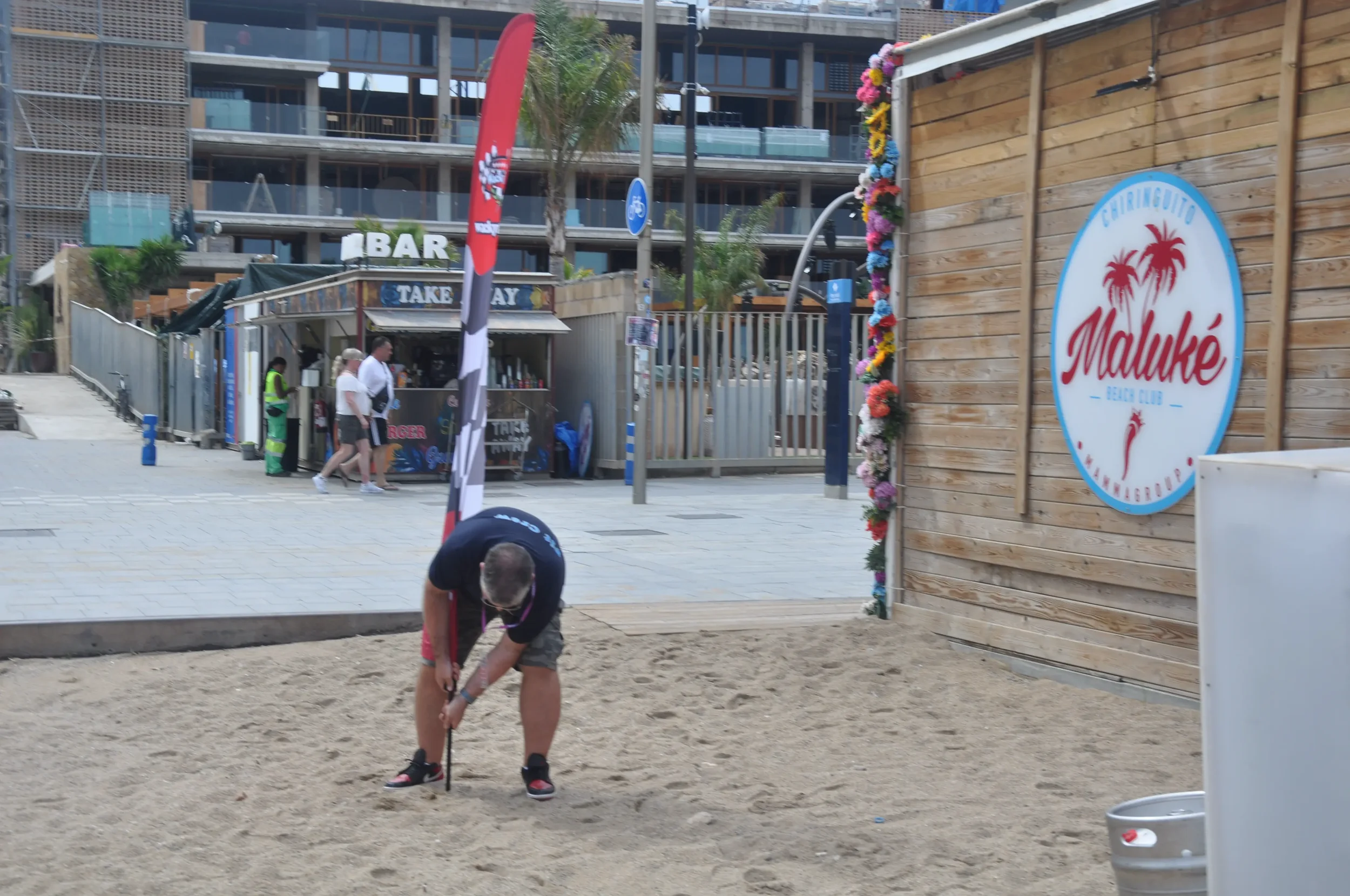 A man in a black t-shirt and shorts is bent over on a sandy beach area, holding a golf club. There is a wooden wall with a sign reading "Maluké Beach Club" decorated with flowers, and a tall flag in the background. People are walking in the backgroun