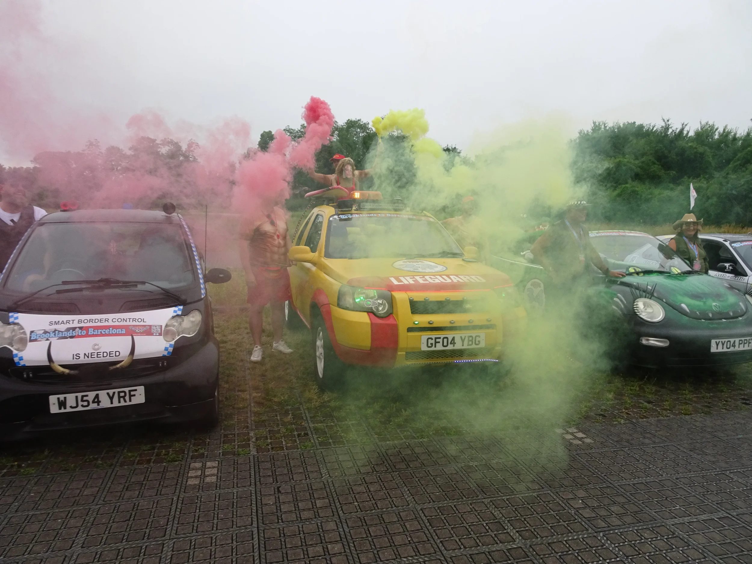 Cars decorated as police, lifeguard, and security with colored smoke at outdoor event, with people dressed in casual and theme outfits.