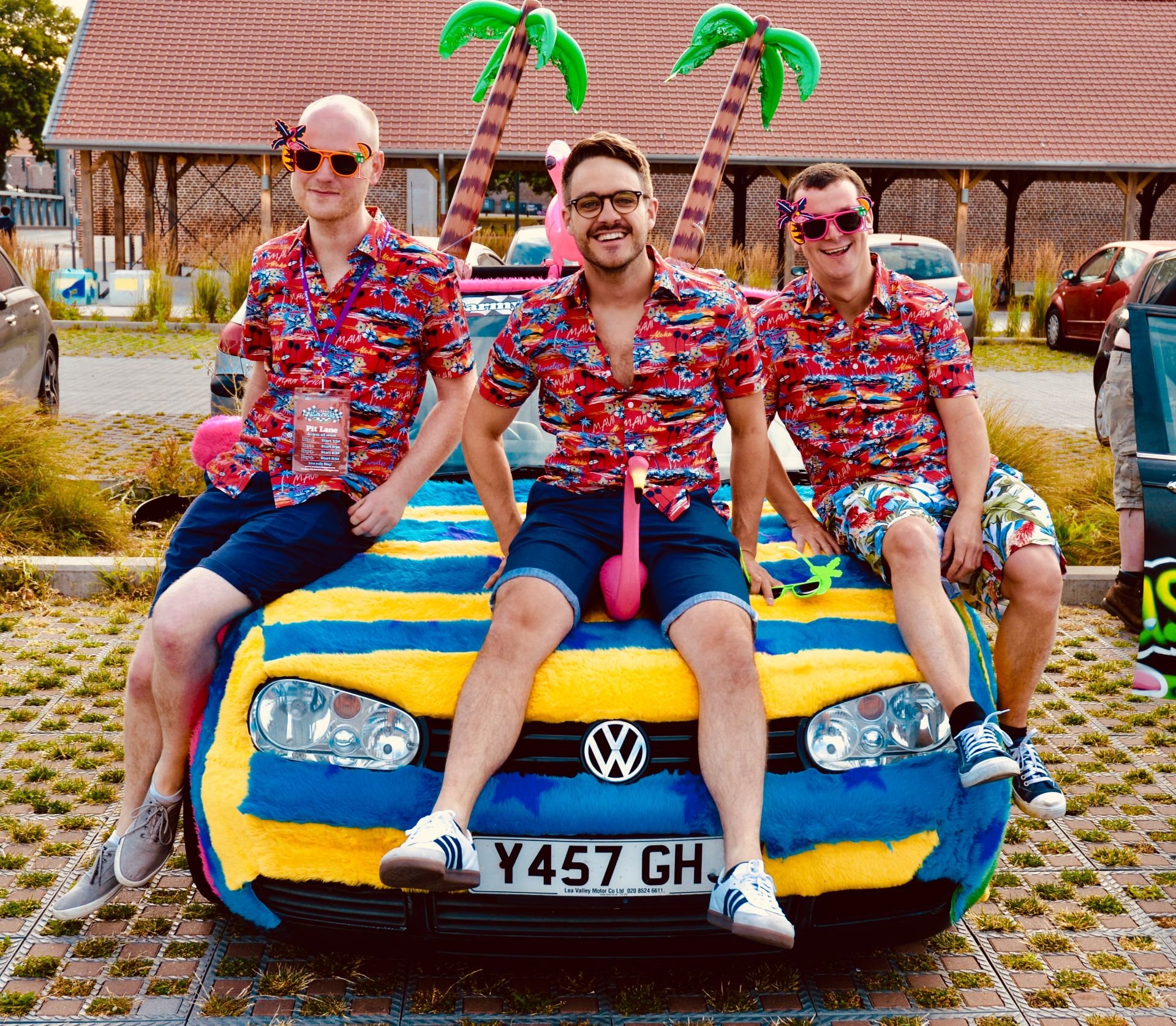 Three men in tropical shirts sitting on the hood of a decorated Volkswagen car at a summer outdoor event. The car is covered with a large, colorful towel and carnival-style accessories, including inflatable palm trees and pink flamingos. All three ar