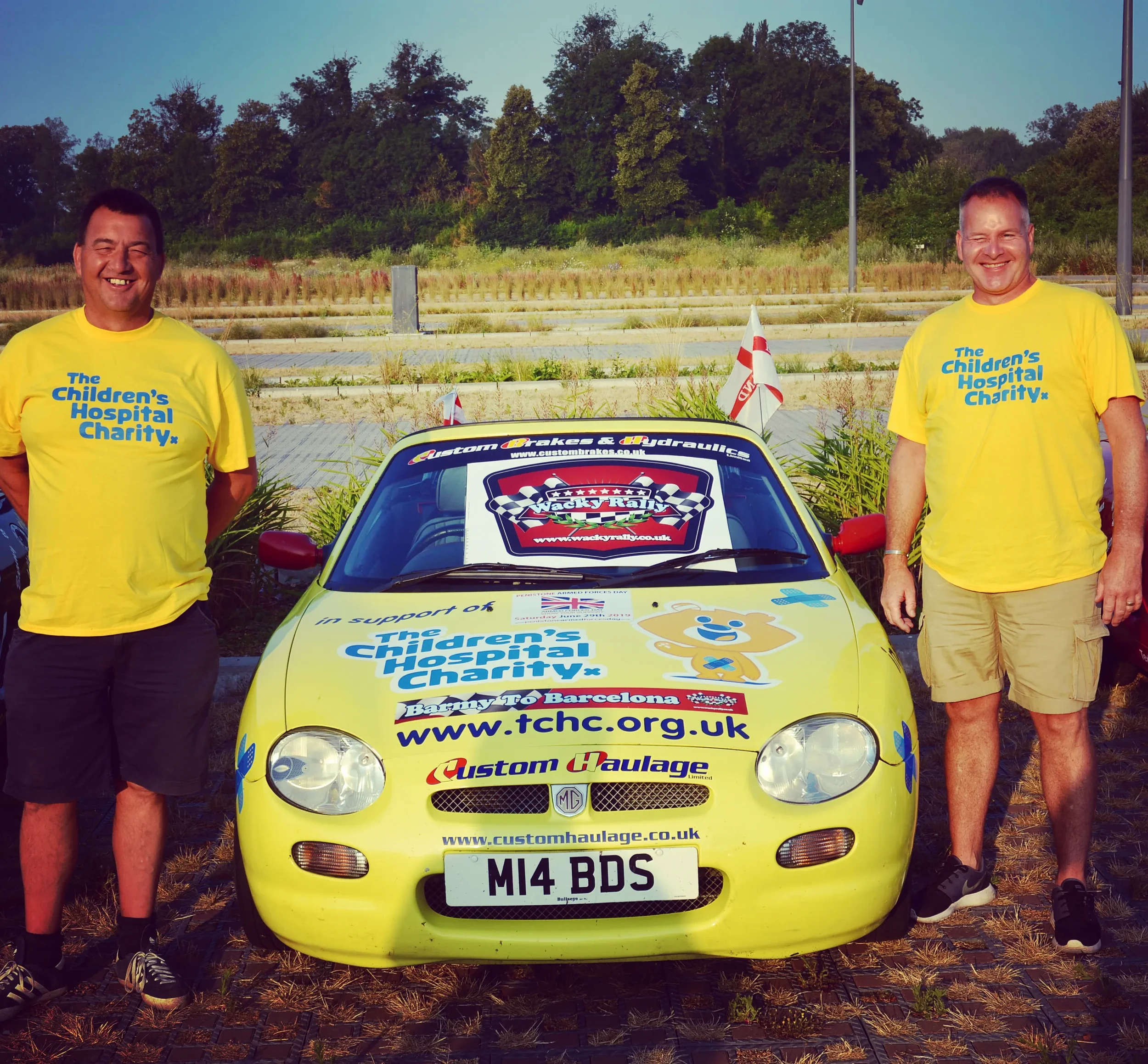 Two men wearing yellow t-shirts with blue text standing on either side of a yellow race car. The car displays logos and web addresses supporting The Children's Hospital Charity, with flags attached, parked in an outdoor area with trees and a partly c