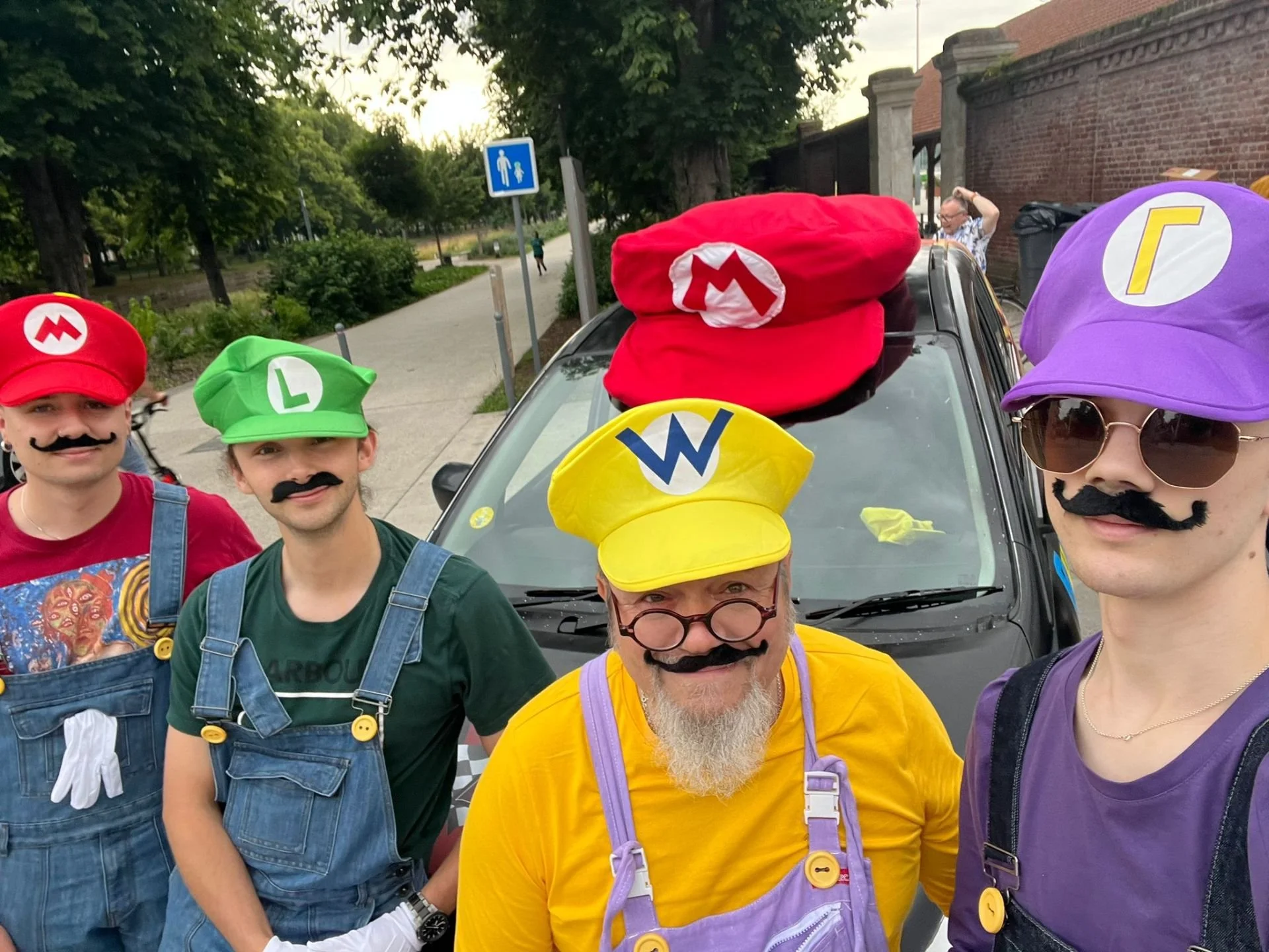 Five people dressed as Mario characters from the Super Mario video game, wearing colorful hats with initials, fake mustaches, and overalls, posing for a photo outdoors.