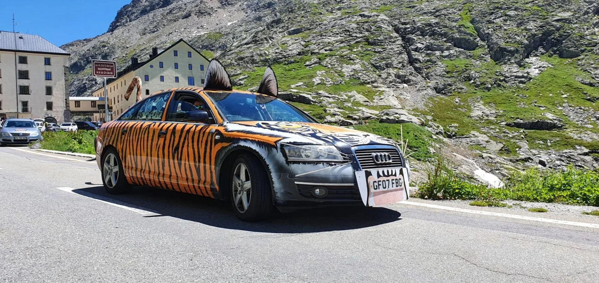 A car with tiger striping and animal features, including cat ears and a painted face, parked on a mountain roadside.