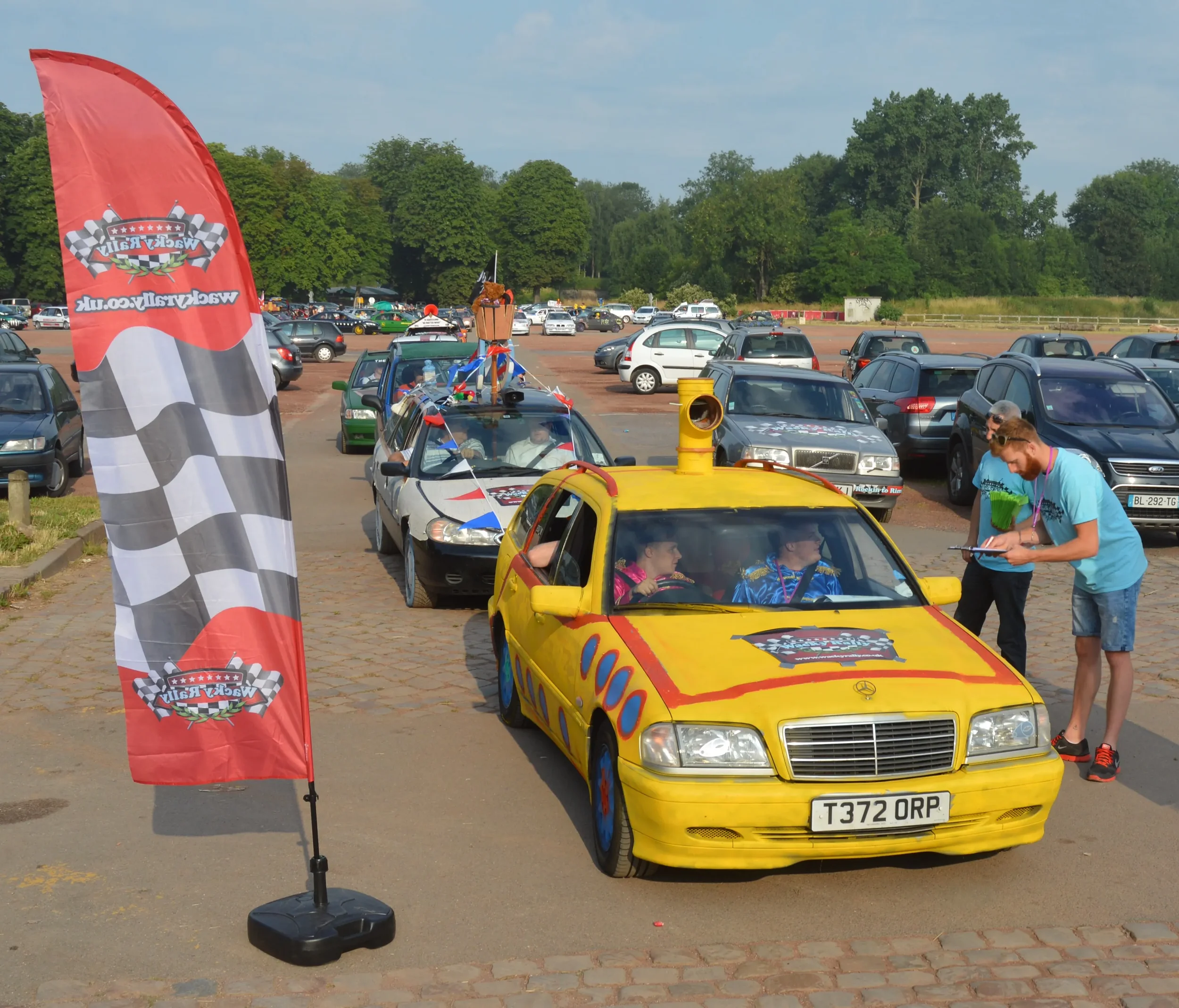 A yellow car decorated with a loudspeaker on the roof, parked in a lot with other cars and a red checkered flag banner nearby. Two people are inside the yellow car, and two individuals are standing outside, one taking notes.
