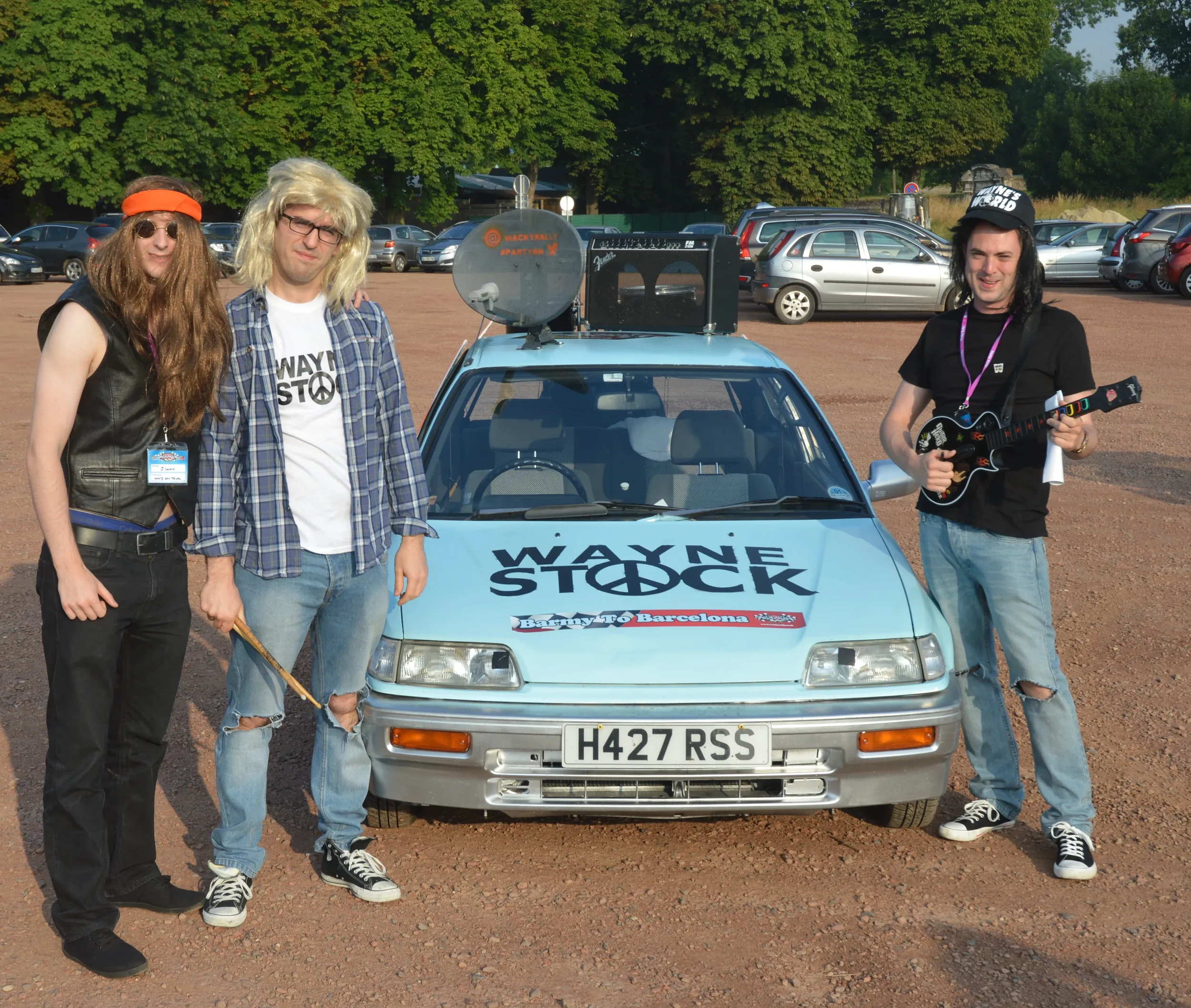Four people dressed in 1980s rock band costumes standing next to a small blue car with a 'Wayne Stock' sticker on the hood, in a parking lot surrounded by trees.