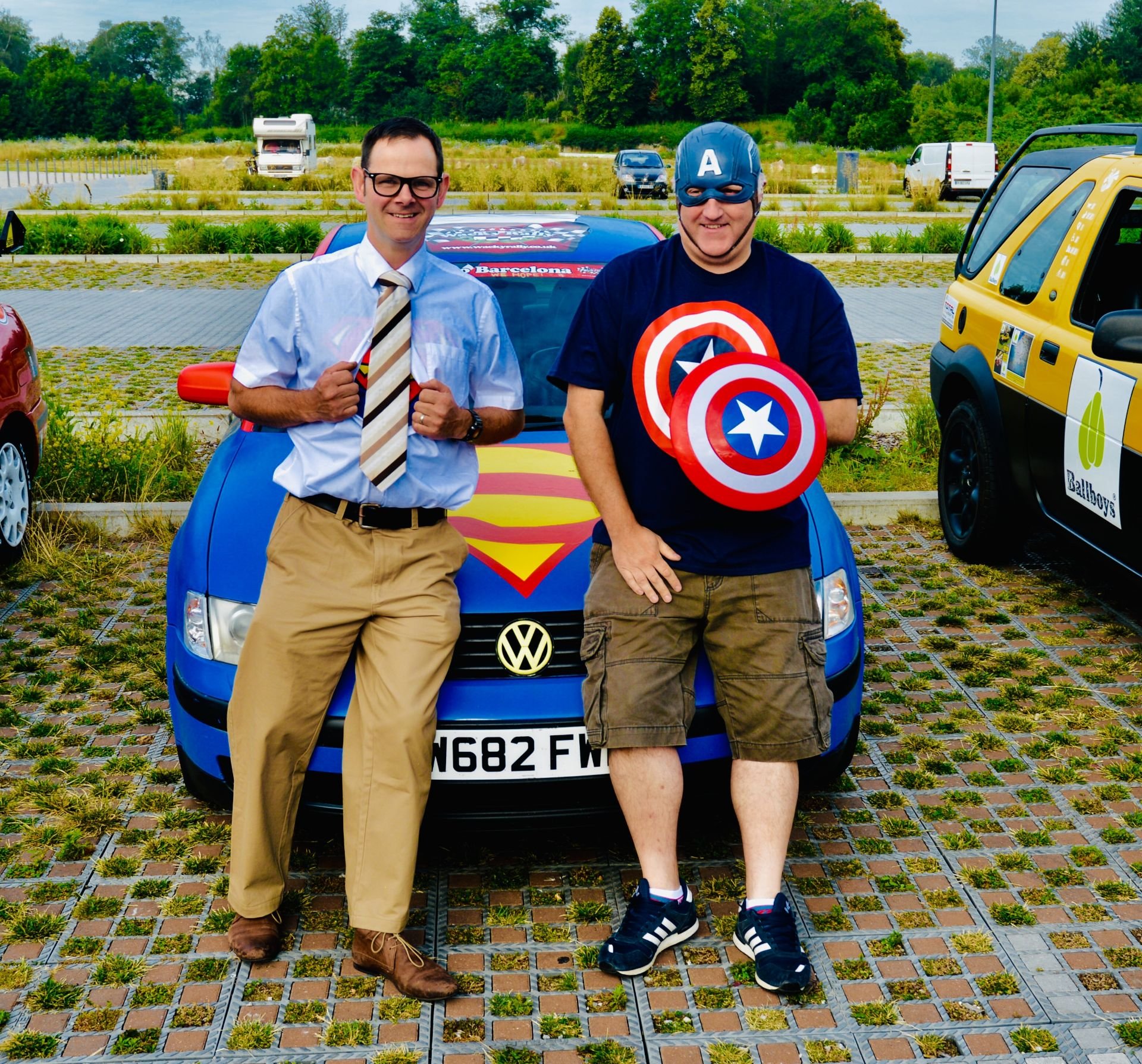 Two men dressed in superhero costumes, one with a Superman logo and the other with Captain America shields, standing in front of a Volkswagen car decorated with superhero themes, in a parking lot with other cars and greenery in the background.