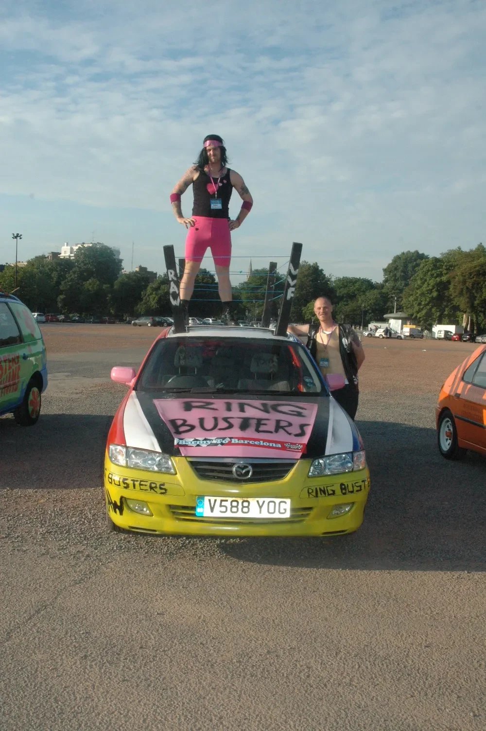 Person dressed in pink and black standing on the roof of a decorated car with 'Ring Busters' signs, with another person standing beside it, in a parking lot with trees in the background.