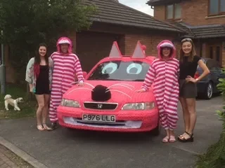Four women standing in front of a pink car with cartoon eyes and antennae, two of them wearing pink and white striped outfits and helmets, while the other two are dressed casually. There is a dog in the background on the lawn, and houses and trees in