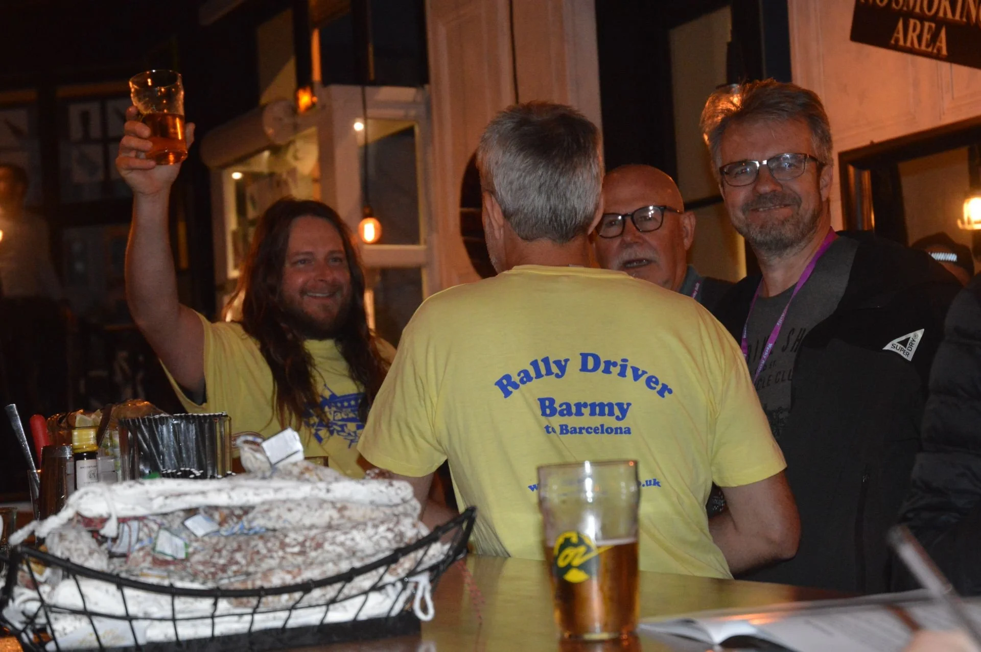 Group of men socializing inside a bar, one wearing a yellow T-shirt with 'Rally Driver Barmy Barcelona' written on the back, holding up a glass of beer, with others smiling and talking.