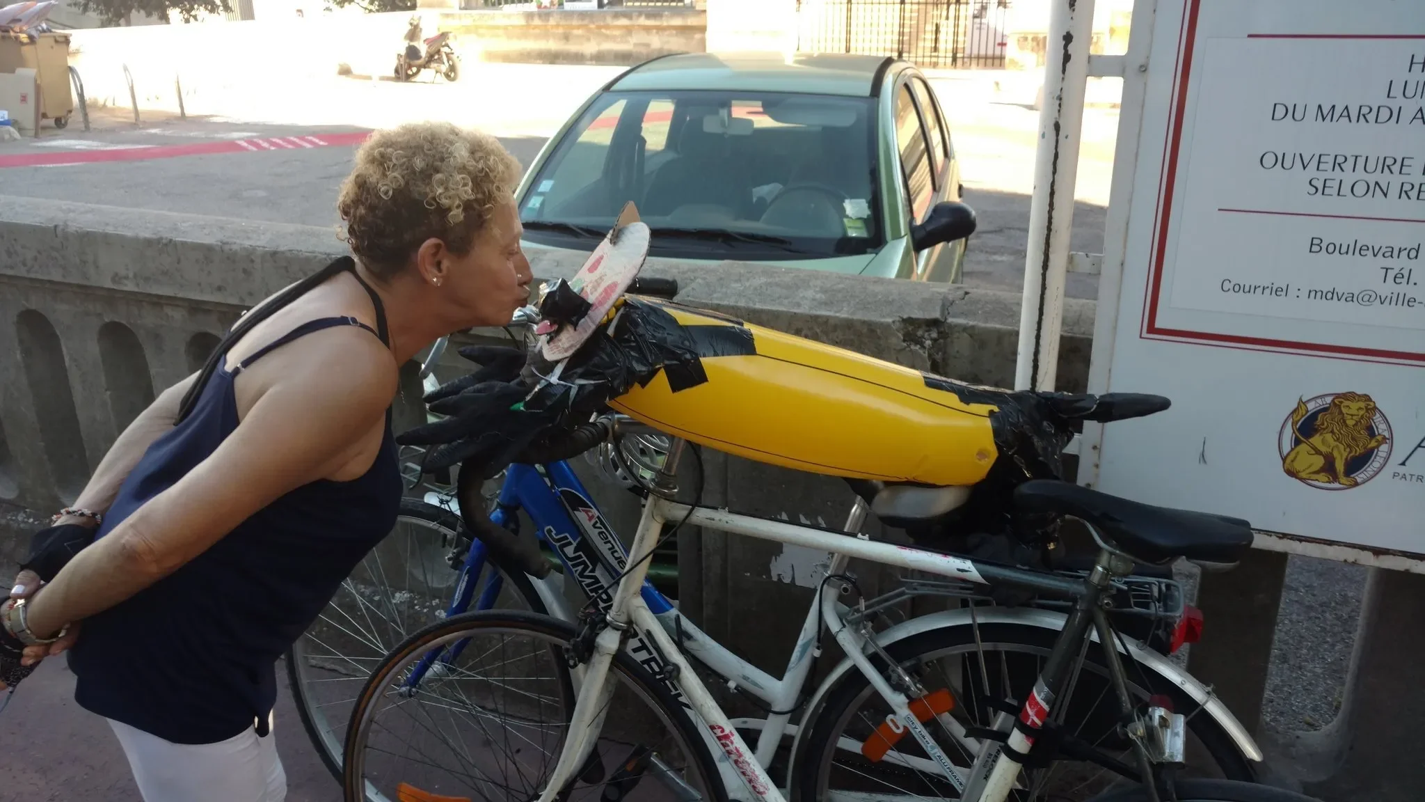 Woman leaning over a bicycle with a yellow cargo container attached, parked on a sidewalk beside a stone wall, with a car and a motorcycle in the background.