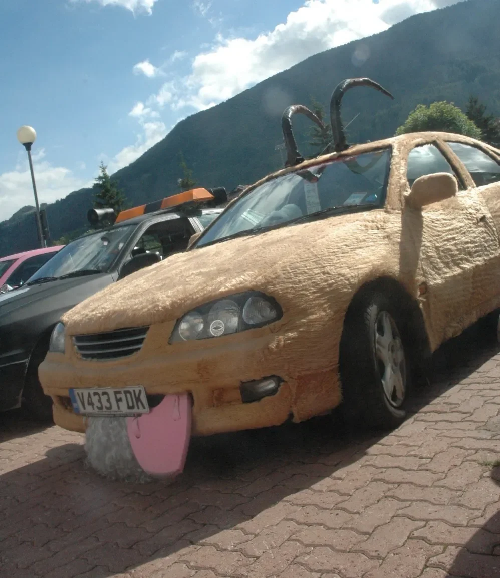 A car covered in a realistic-looking sheep fur costume, with pink tongue and black horns on the roof, parked next to other vehicles in a parking lot with mountains in the background.