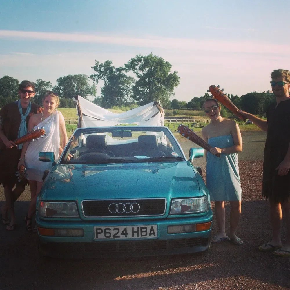 Group of five young adults standing around a teal vintage Audi convertible car, holding inflatable guitars, outdoors on a sunny day with trees and open field in the background.