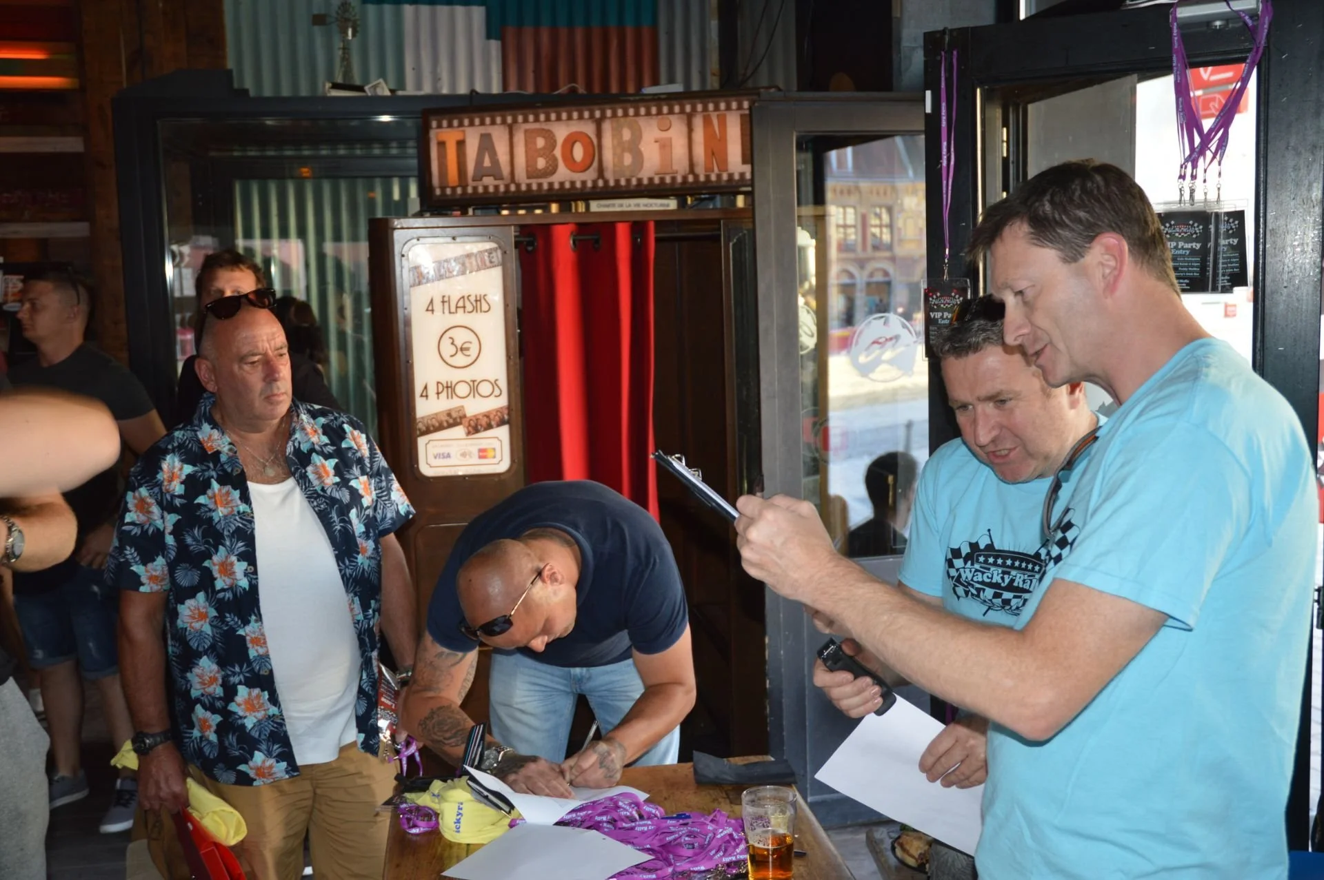 A group of people at an outdoor event, with a man wearing sunglasses and a floral shirt standing next to a man signing something on a table. Two men are looking at a device or paper, one of whom is wearing a blue t-shirt with the logo "Wacky R". Ther
