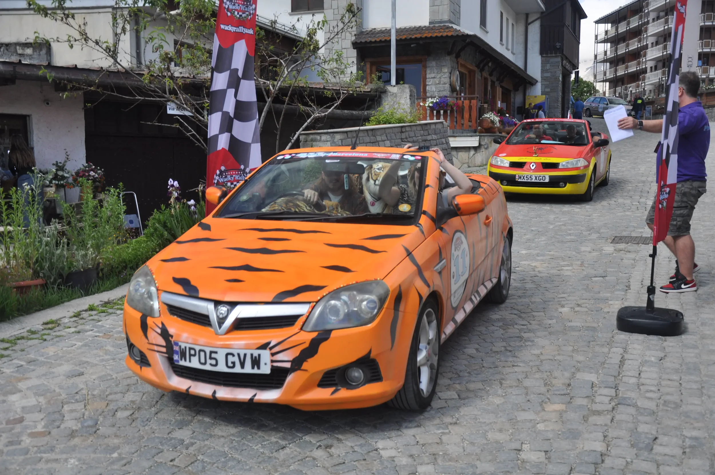 A parade with cars decorated like a tiger and a Ferrari at a race event, with flags and people involved in the event.