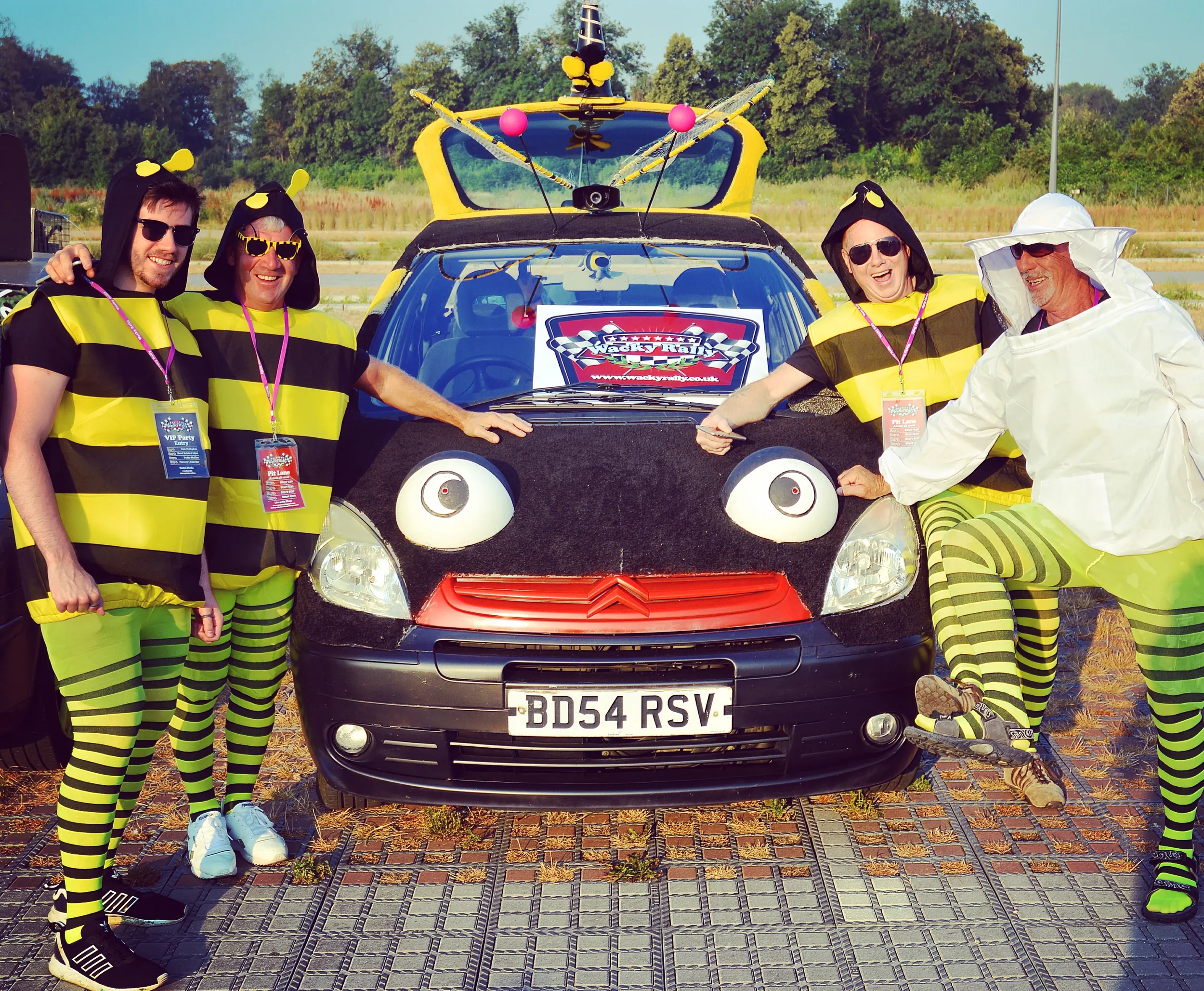 A group of five people dressed in bee and hive costumes, standing around a car with a face design and bee decorations. They are outdoors on a sunny day, smiling and posing for the photo.