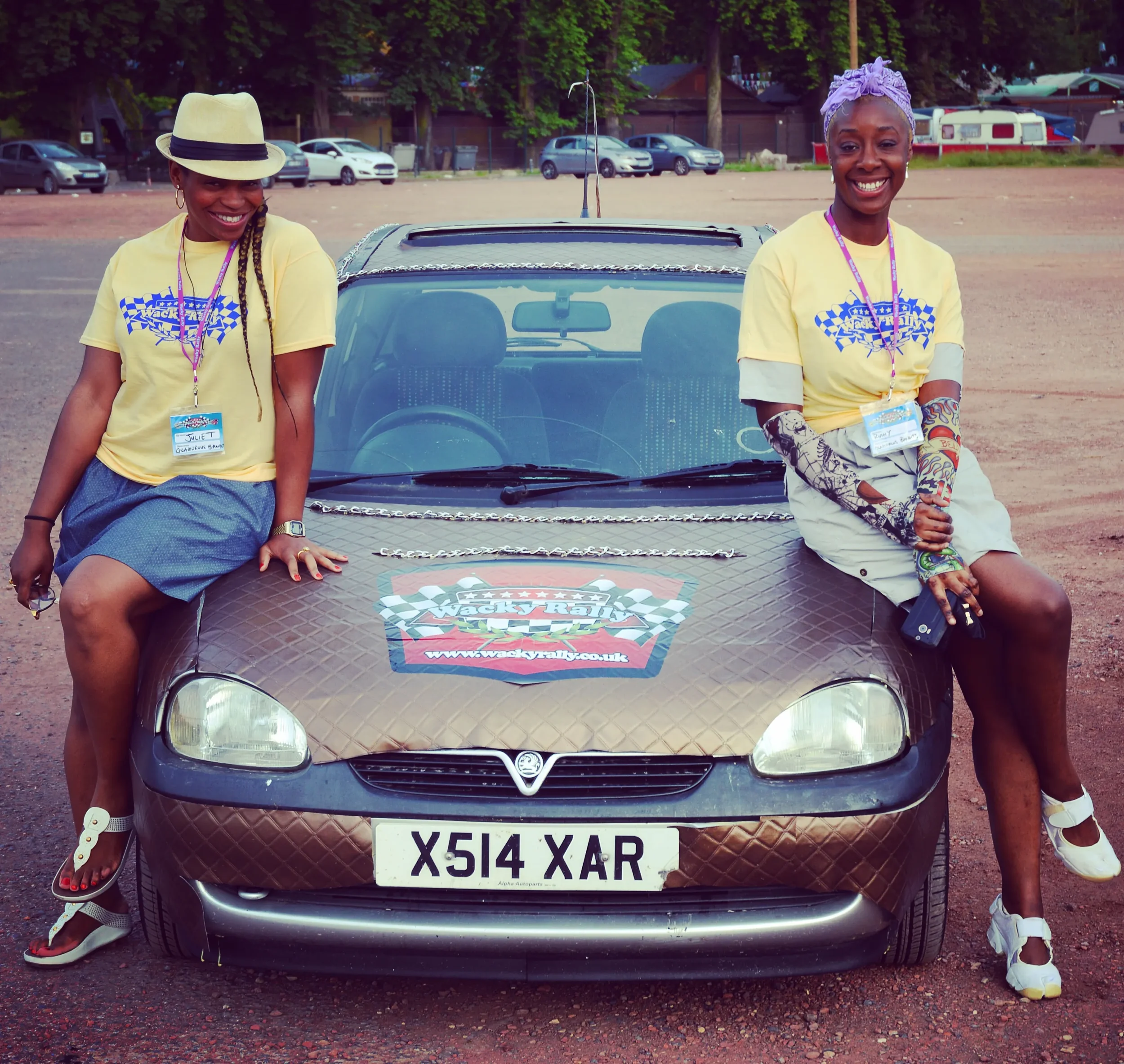 Two women with name tags posing beside a brown Vauxhall car with racing stickers, on a dirt lot with parked cars and trees in the background.