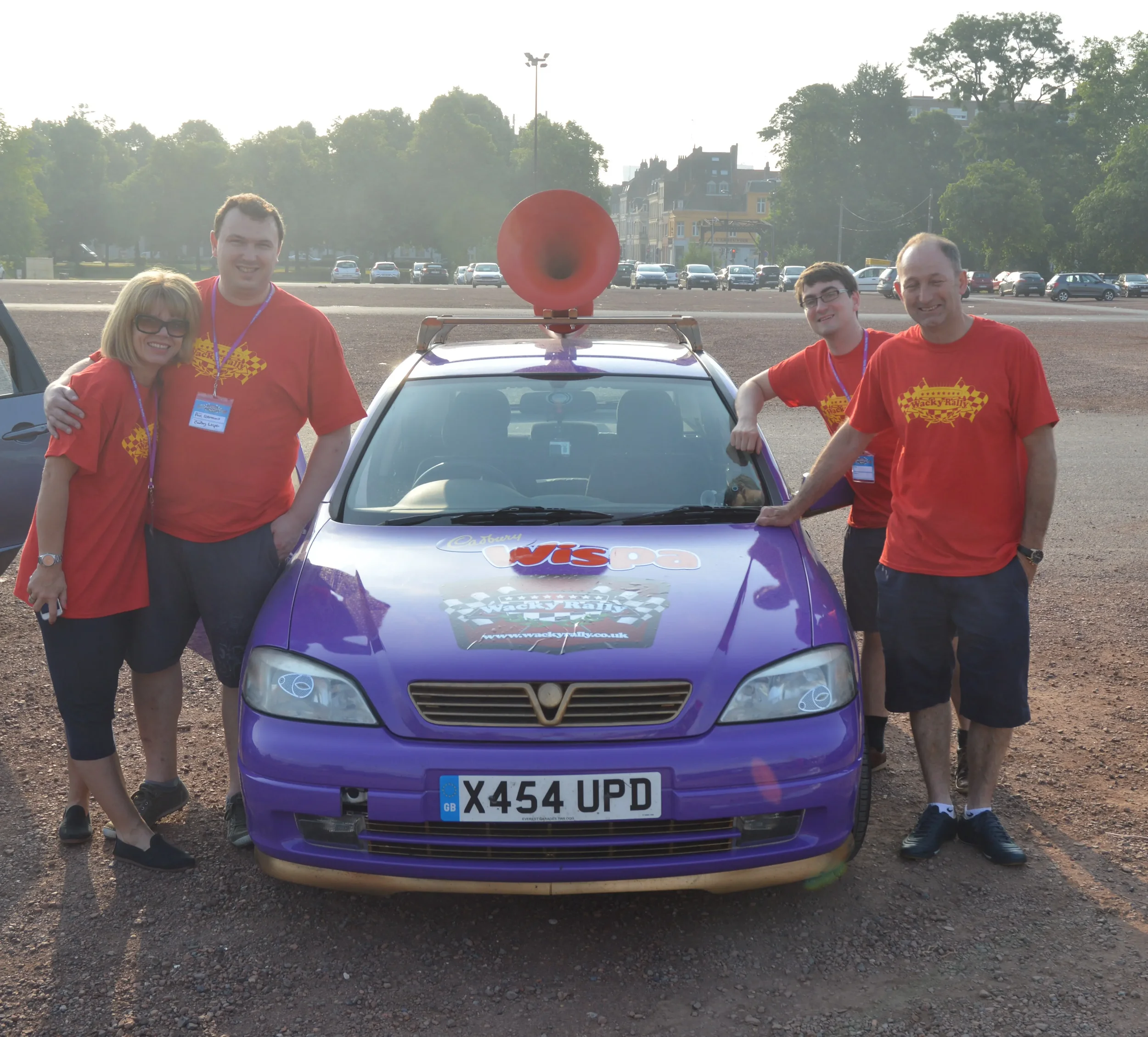 Group of four people standing around a purple Vauxhall Astra car with a large red megaphone on its roof, in an outdoor parking area during daytime, all wearing red T-shirts with a racing checkered flag design.