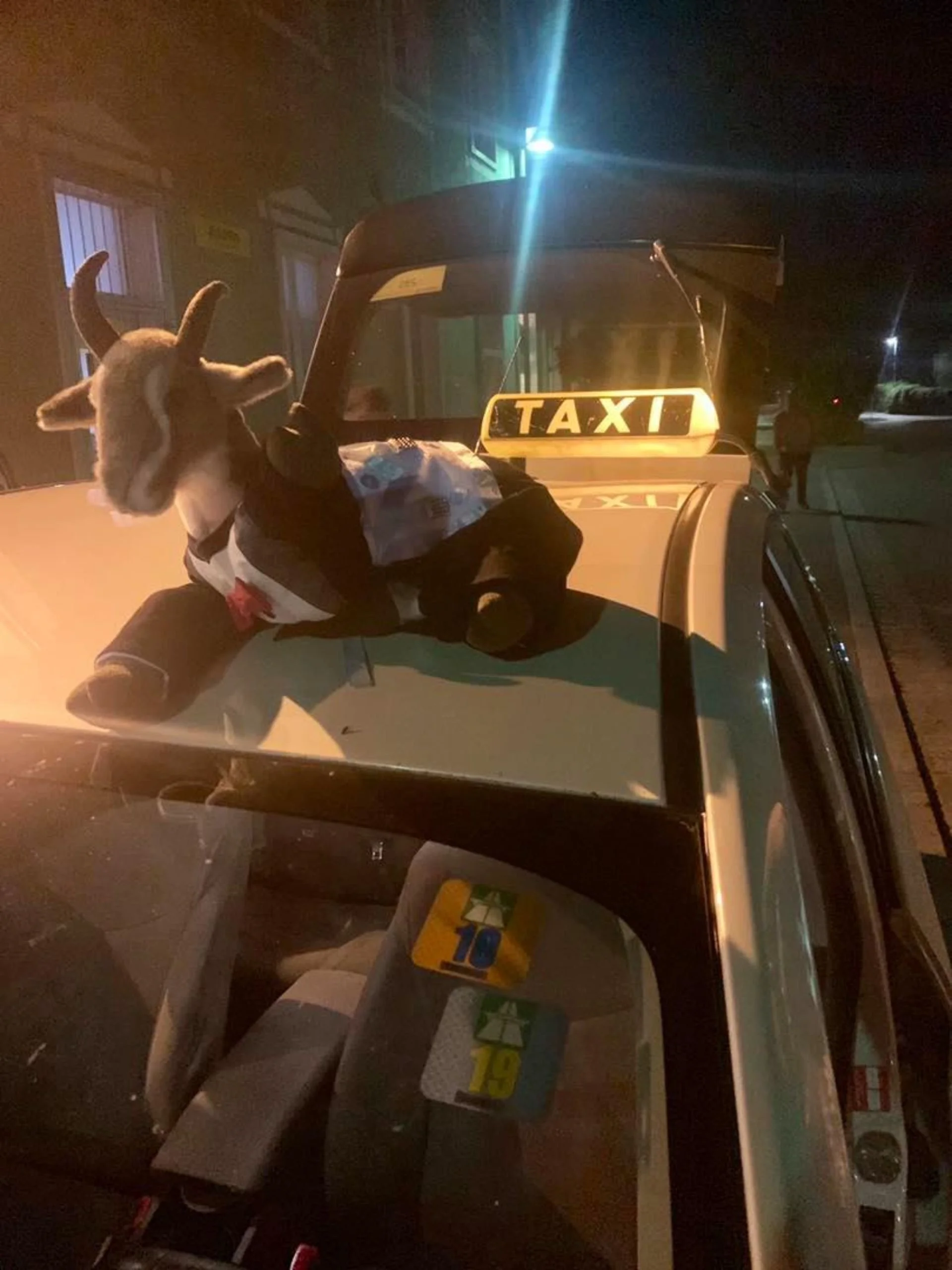 A stuffed animal goat dressed as a taxi driver sitting on the roof of a taxi car at night, with a city street in the background.