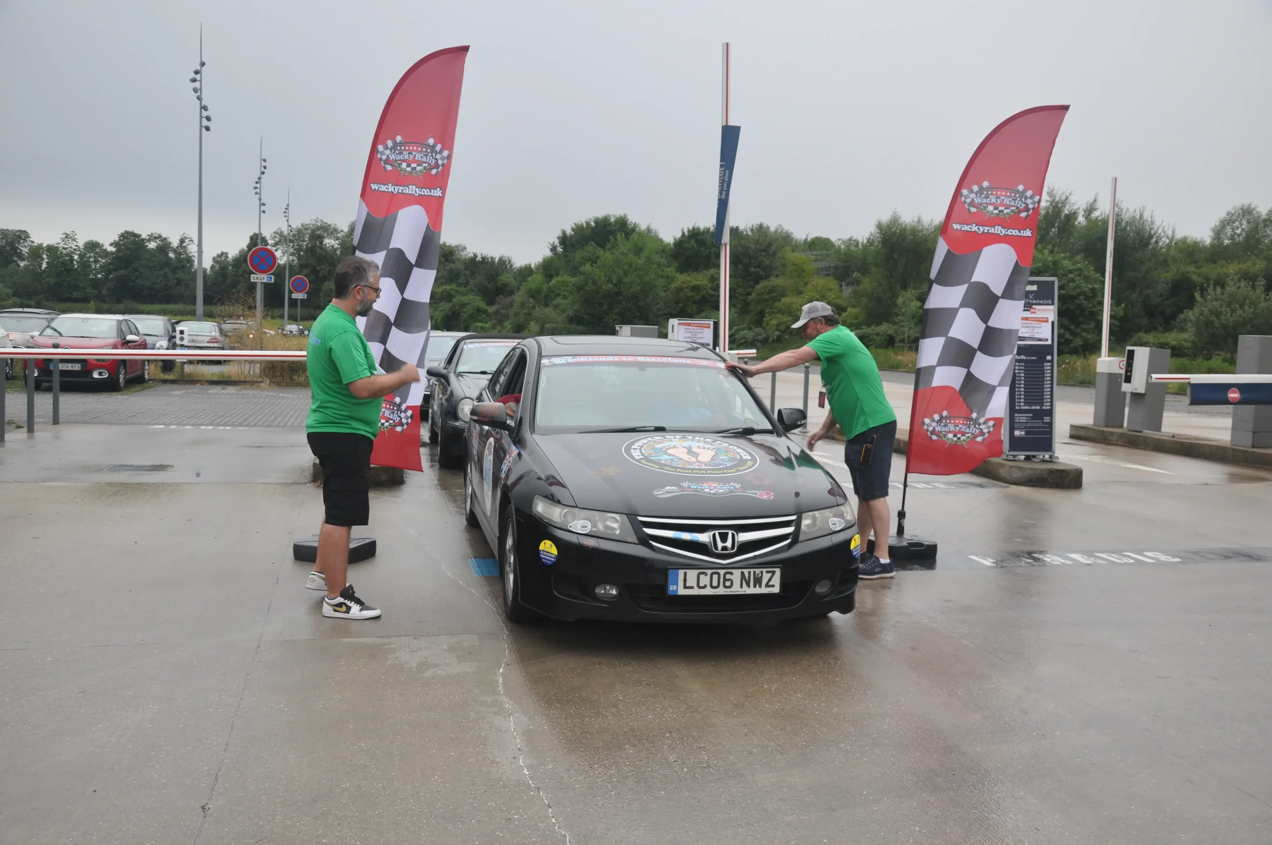 A black Honda car at a rally finish line with three men in green shirts nearby, two holding flags with checkered patterns.
