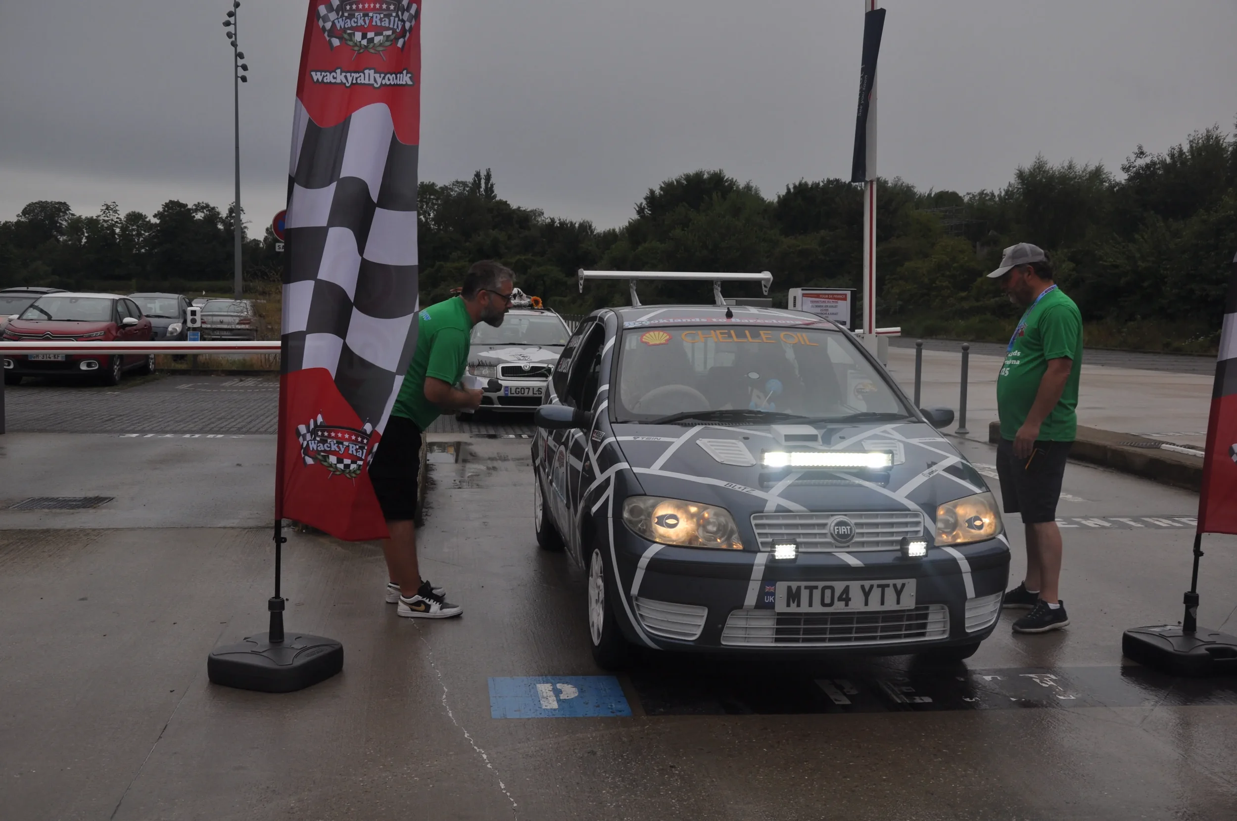 A rally car with the license plate 'MT04 YTY' parked under a tent with two men in green shirts inspecting the vehicle. The car has sponsorship decals including 'Shell Oil' and features additional rally lights. The scene is set on a wet day with overc