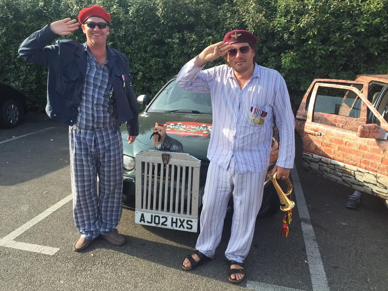 Two men in pajamas and military-style berets dressed as soldiers standing in a parking lot, saluting. One man is holding a gold whistle, and the other is holding a toy gun. There is a small cart with a dog statue in front of a car with the license pl