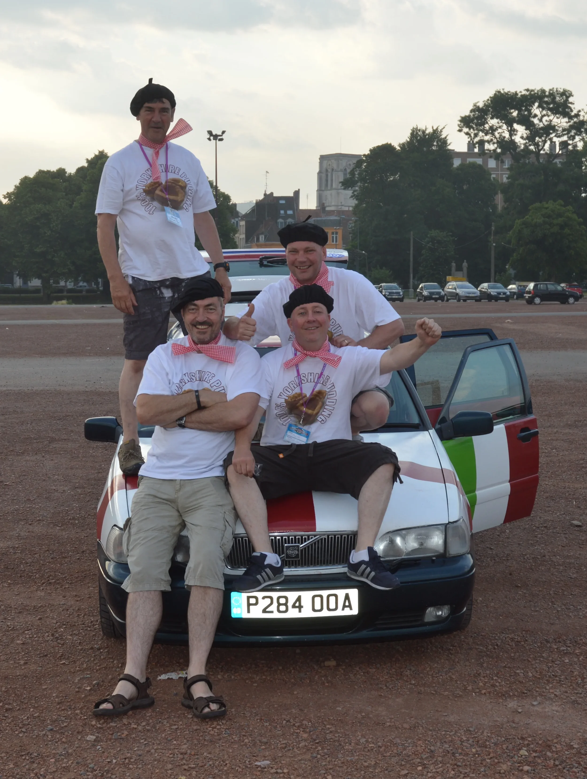 Four men dressed in white shirts with red checkered neckerchiefs and black berets are posing on and around a small car in an outdoor parking lot. The car has a red, white, and green striped pattern on the side. The men are smiling and one is giving a