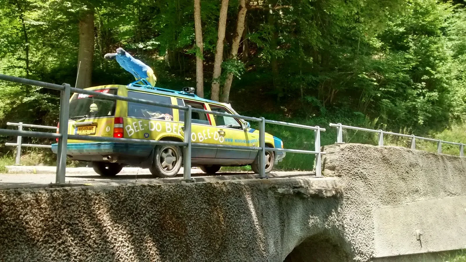 A colorful vehicle decorated with bee-themed design and themes, parked on a bridge next to a wooded area.